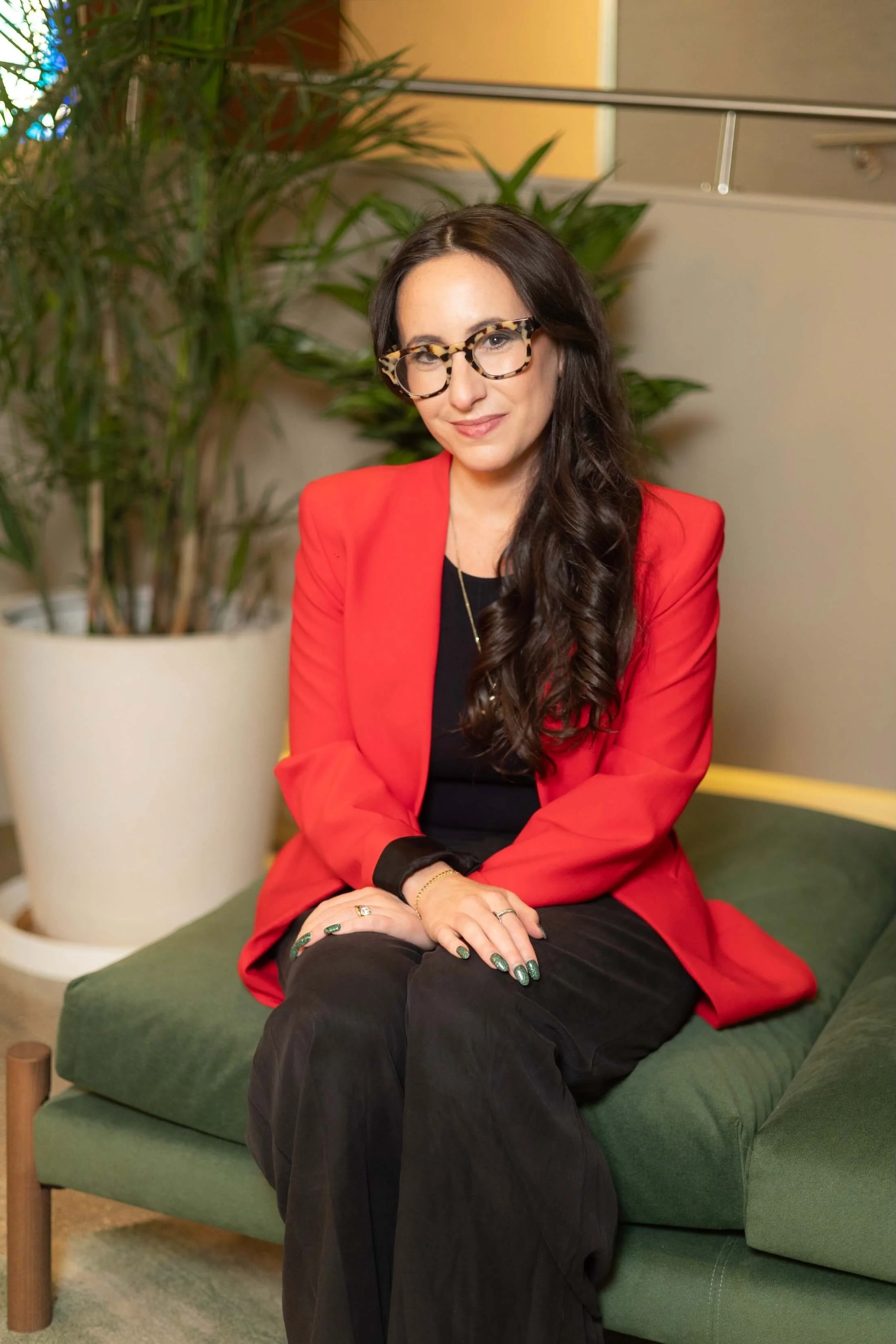 A woman with long dark hair, glasses with a leopard print frame, wearing a red blazer and black pants, sitting on a green cushioned seat in front of a large potted plant with long green leaves, indoors with beige walls.