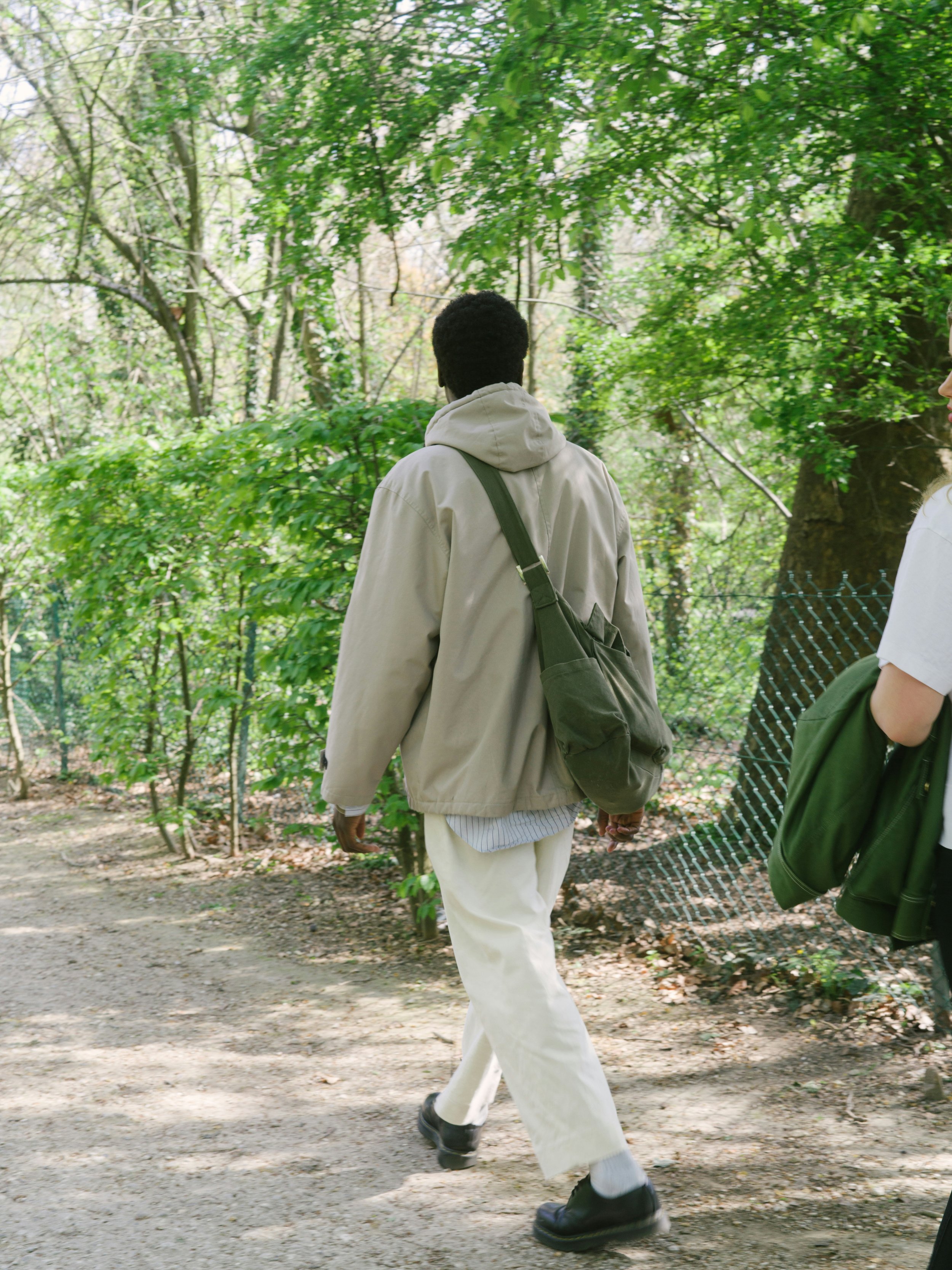 A person walking through a wooded outdoor area, dressed in light-colored pants, a beige jacket, and black shoes, with a backpack on their back, and another person partially visible on the right side.