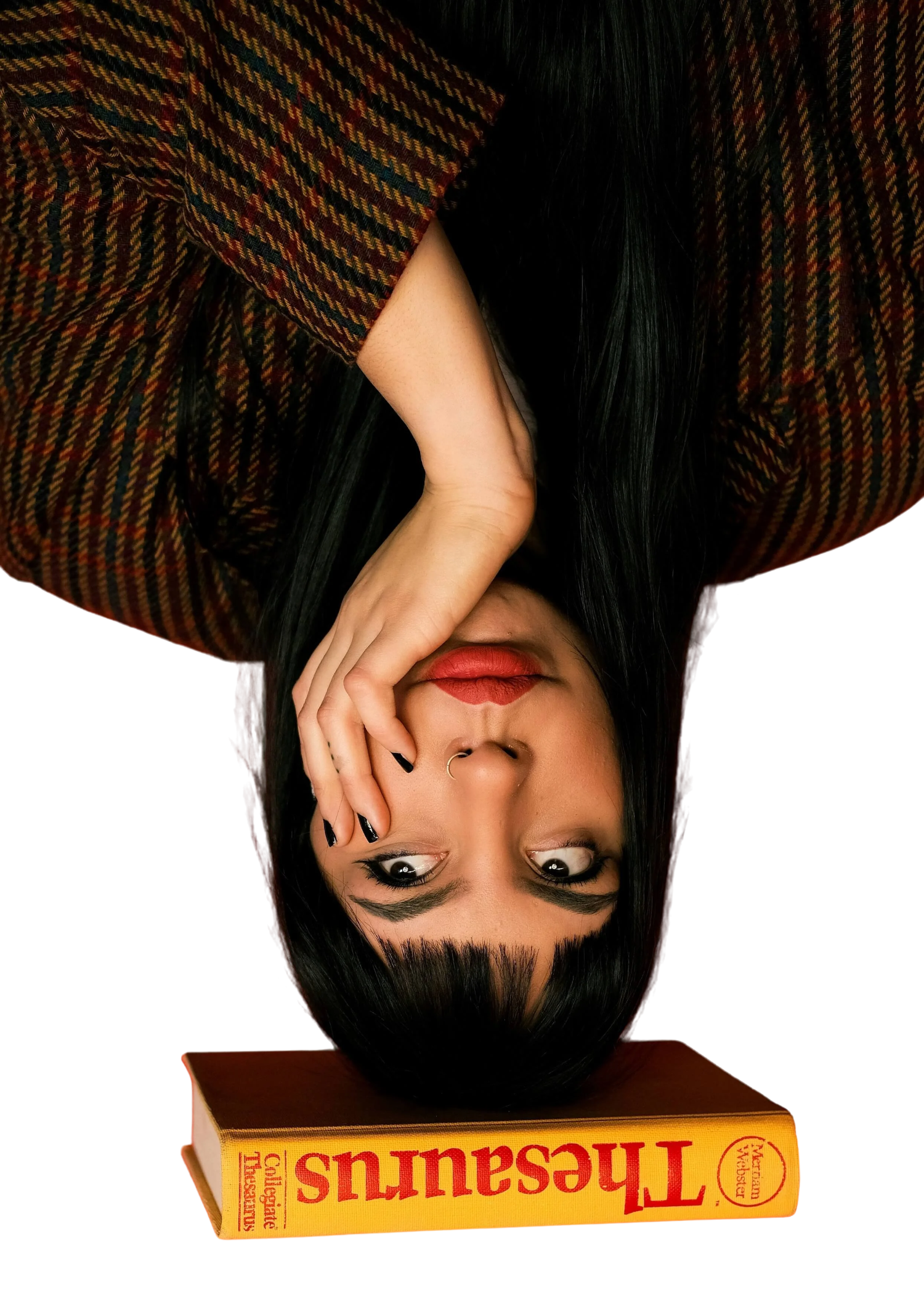 A woman with black hair and makeup laying with her head on a book titled 'Treasaurus: Guide to Geographical Landmarks' by Marian Veblest. She is wearing a plaid shirt and has a nose ring, covering one eye with her hand and looking at the camera.