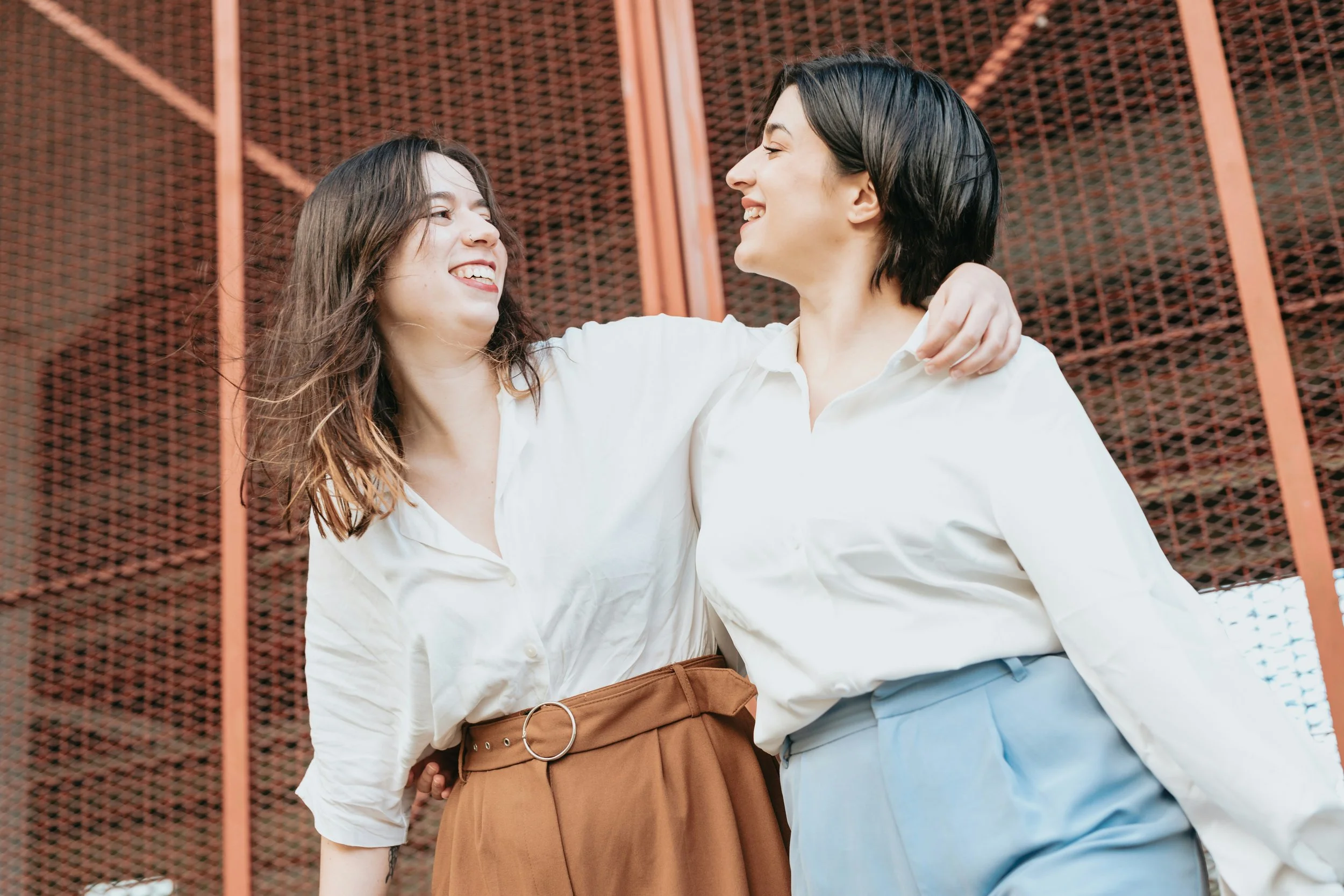 Two women smiling and looking at each other, with one woman having her arm around the other, standing in front of a red metal fence.