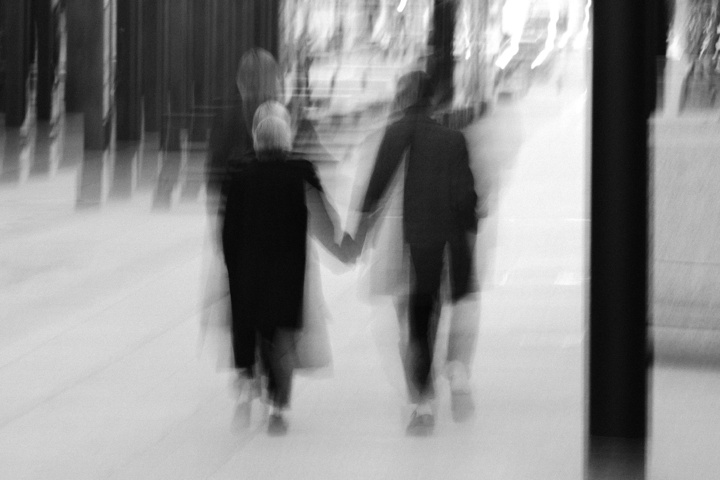 Blurred image of a woman and a man holding hands, walking through an airport terminal with blurred background of walls and windows.