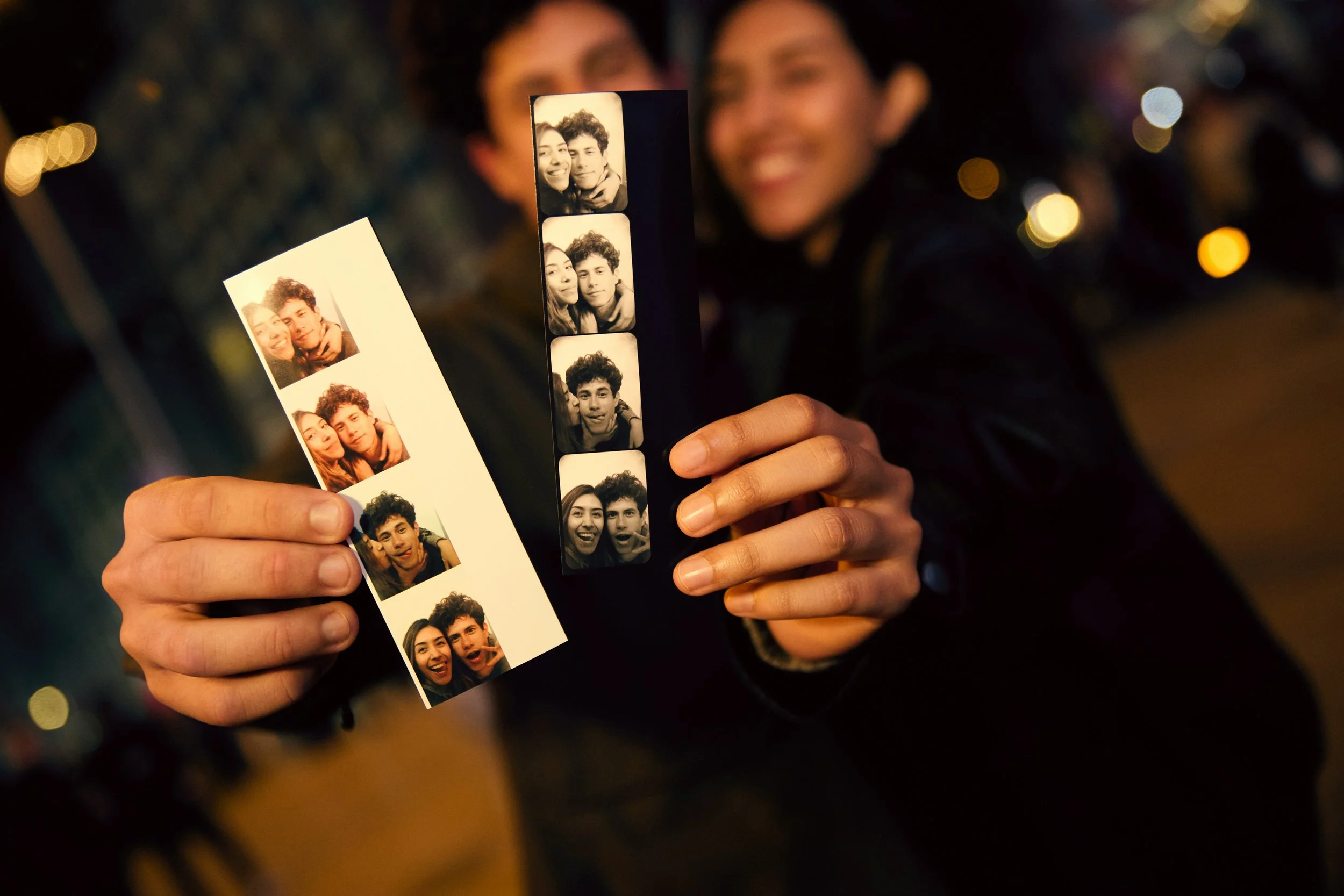 Person holding a strip of photo booth pictures showing a couple smiling and making funny faces, with blurred background of lights and people.