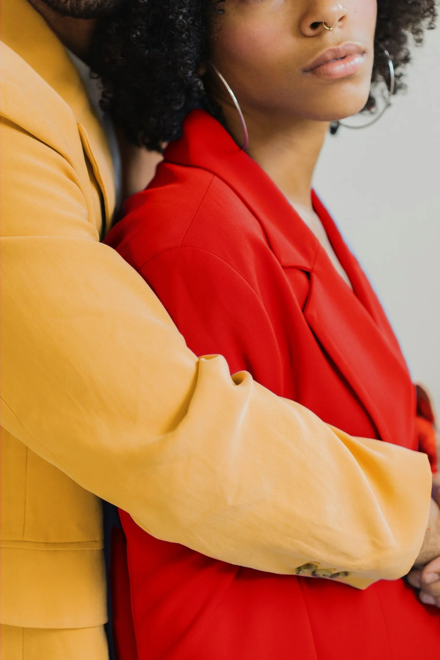 Close-up of two people, one wearing a yellow jacket and the other in a red jacket, showcasing their shoulders and partial faces, with curly hair and gold jewelry, against a plain background.