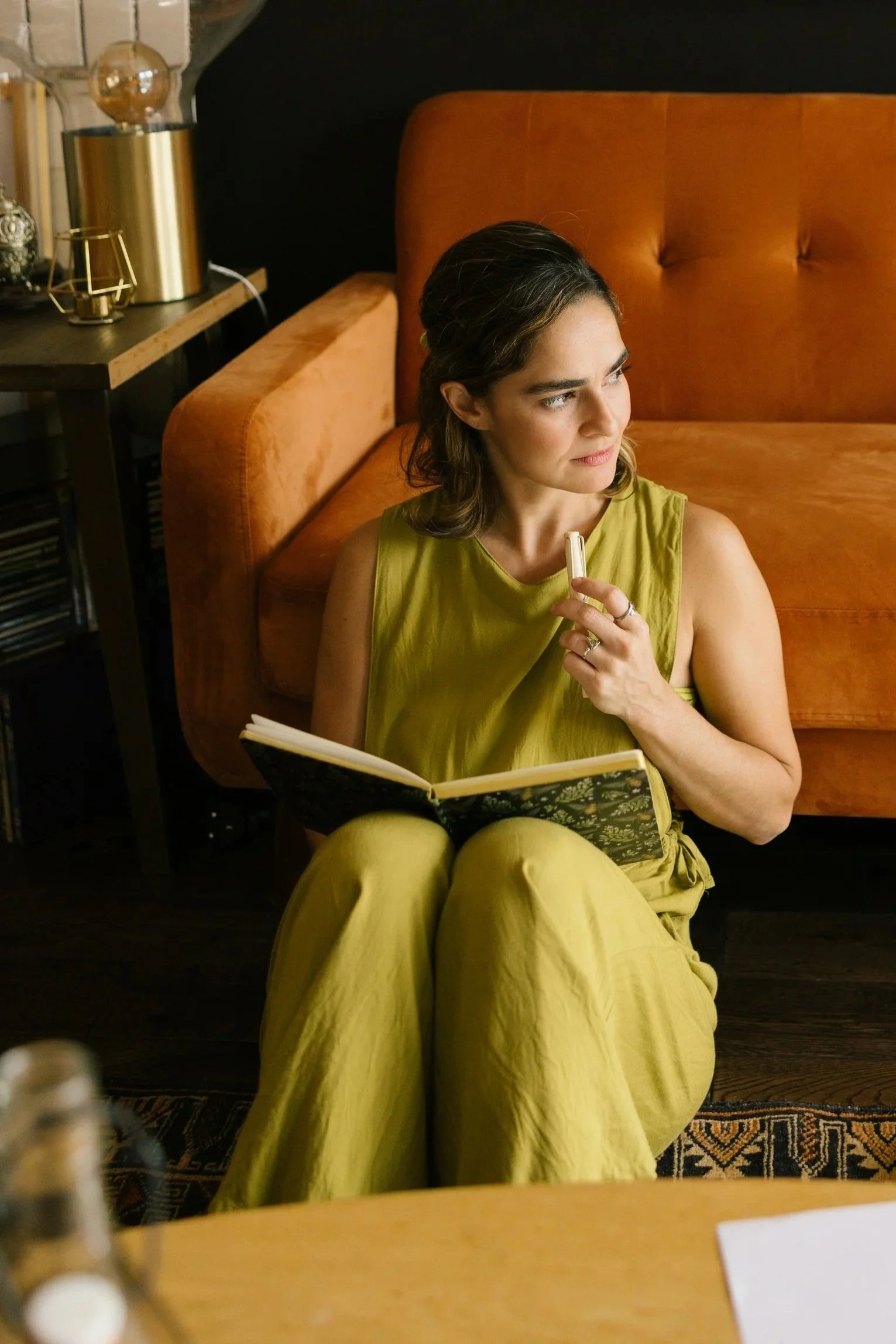 Woman sitting on the floor with a yellow dress, holding a pen and a notebook, in a cozy living room with a brown velvet sofa and decorative lamps.