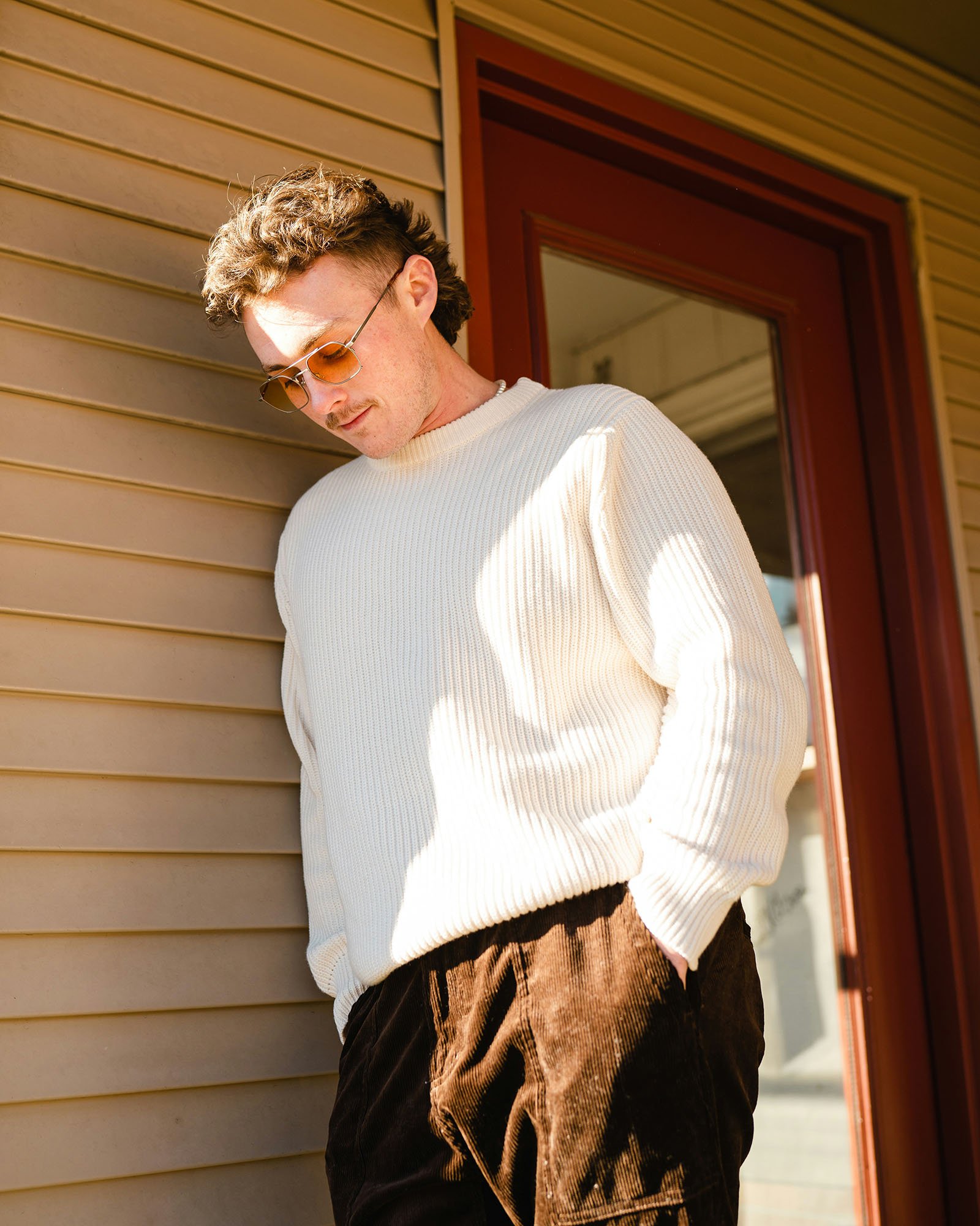 A young man with curly hair and sunglasses wearing a white sweater and brown velvet pants standing beside a house with yellow siding and a red door.