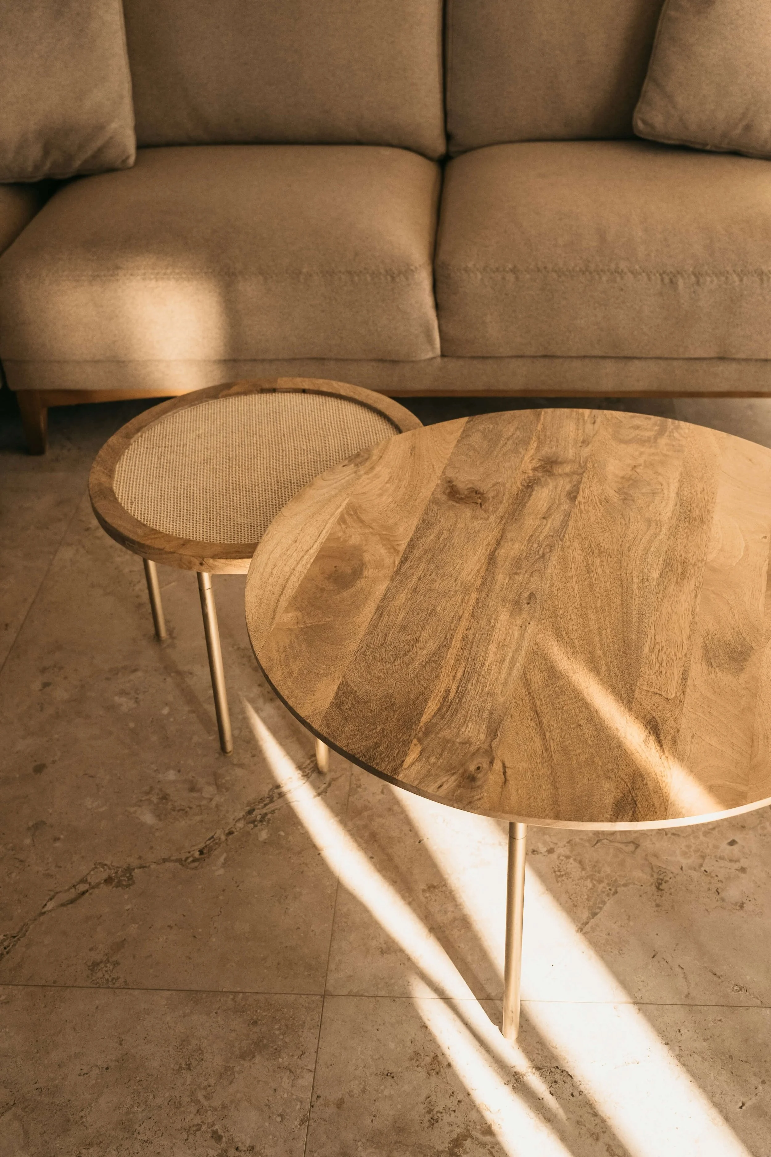 A beige sofa with two cushions and a wooden frame, positioned behind two round wooden tables on a tiled floor with sunlight casting shadows.