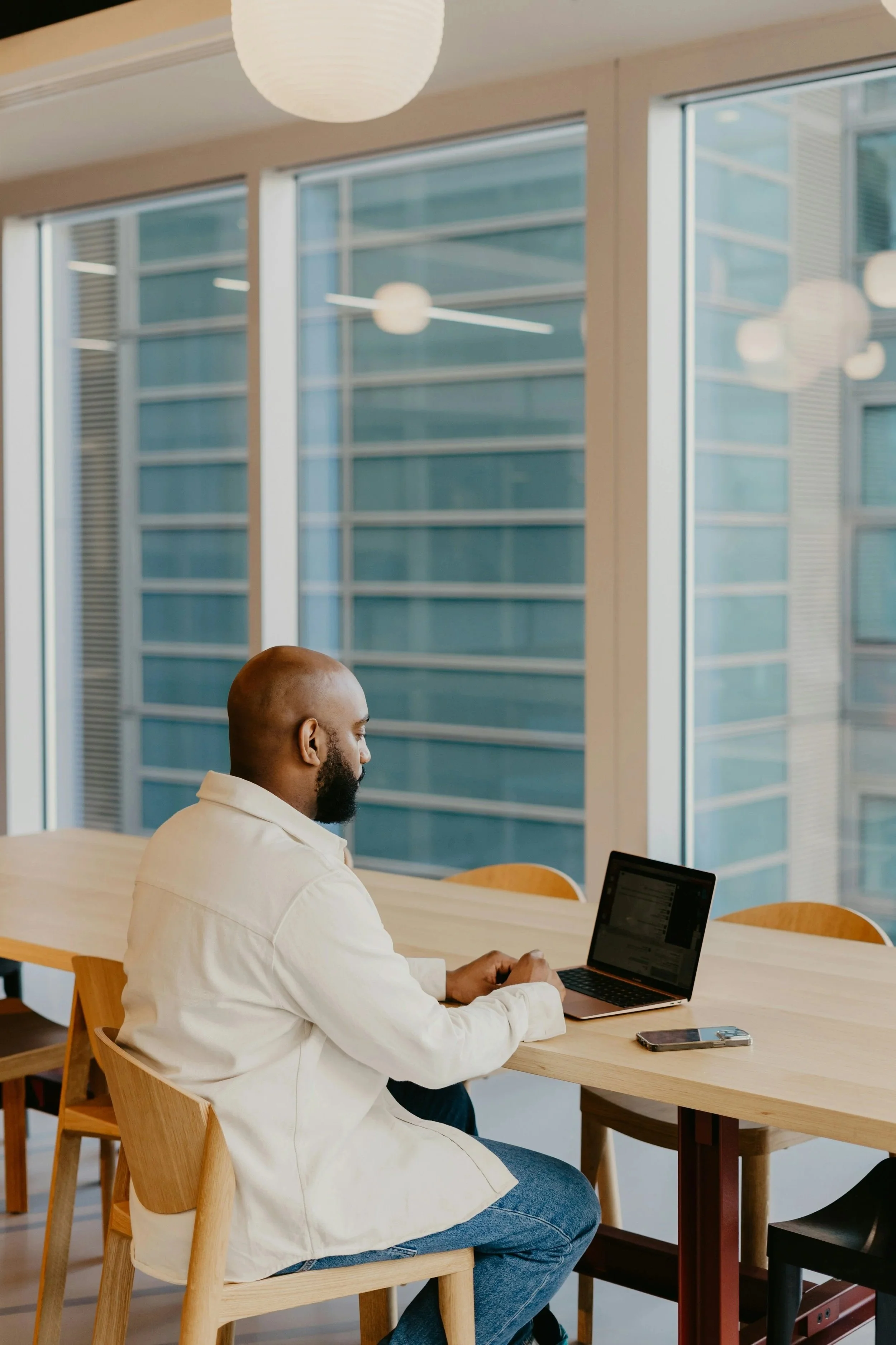 A man with a beard sitting at a wooden table in a room with large windows, using a laptop and having a smartphone nearby.