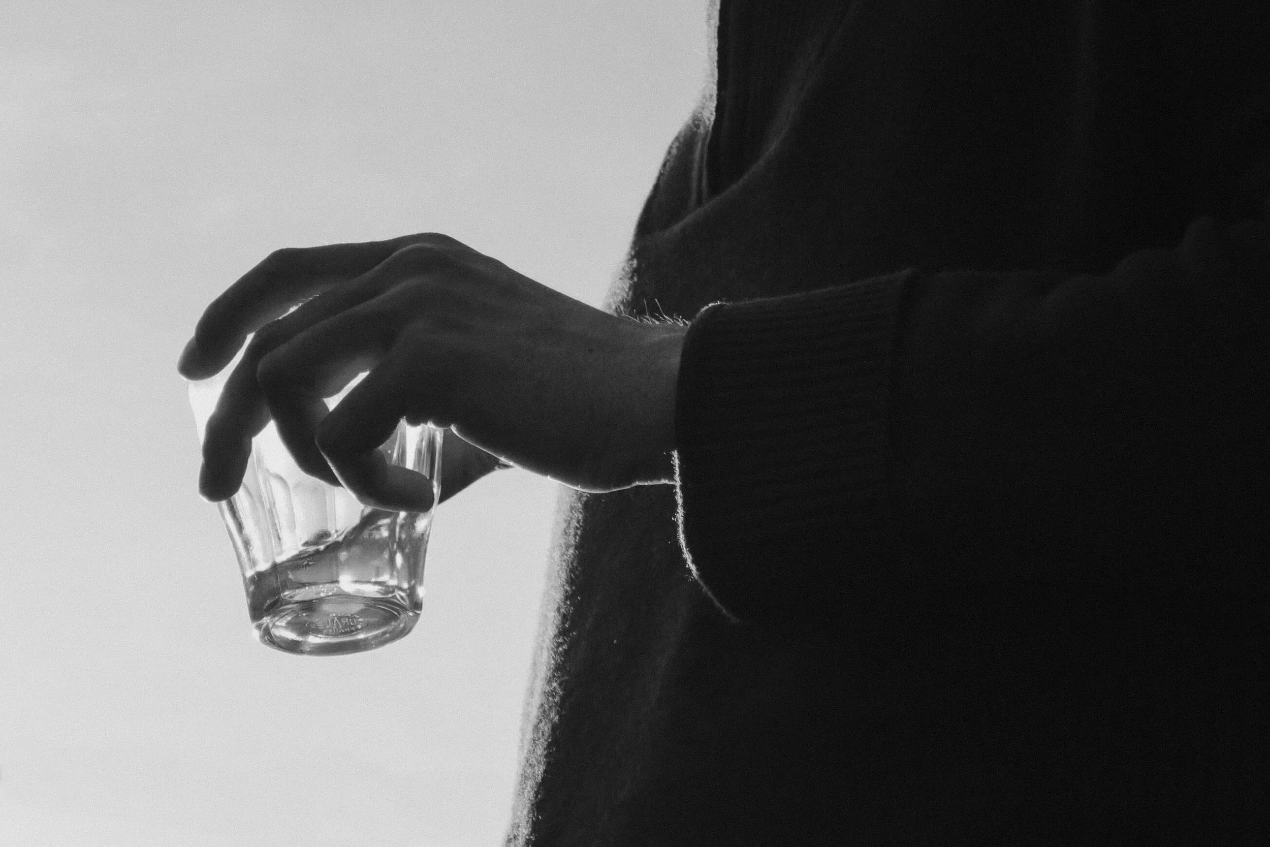 A black and white photo of a person's hand holding a glass of water. The person is wearing a dark sweater and is sitting on grassy ground.