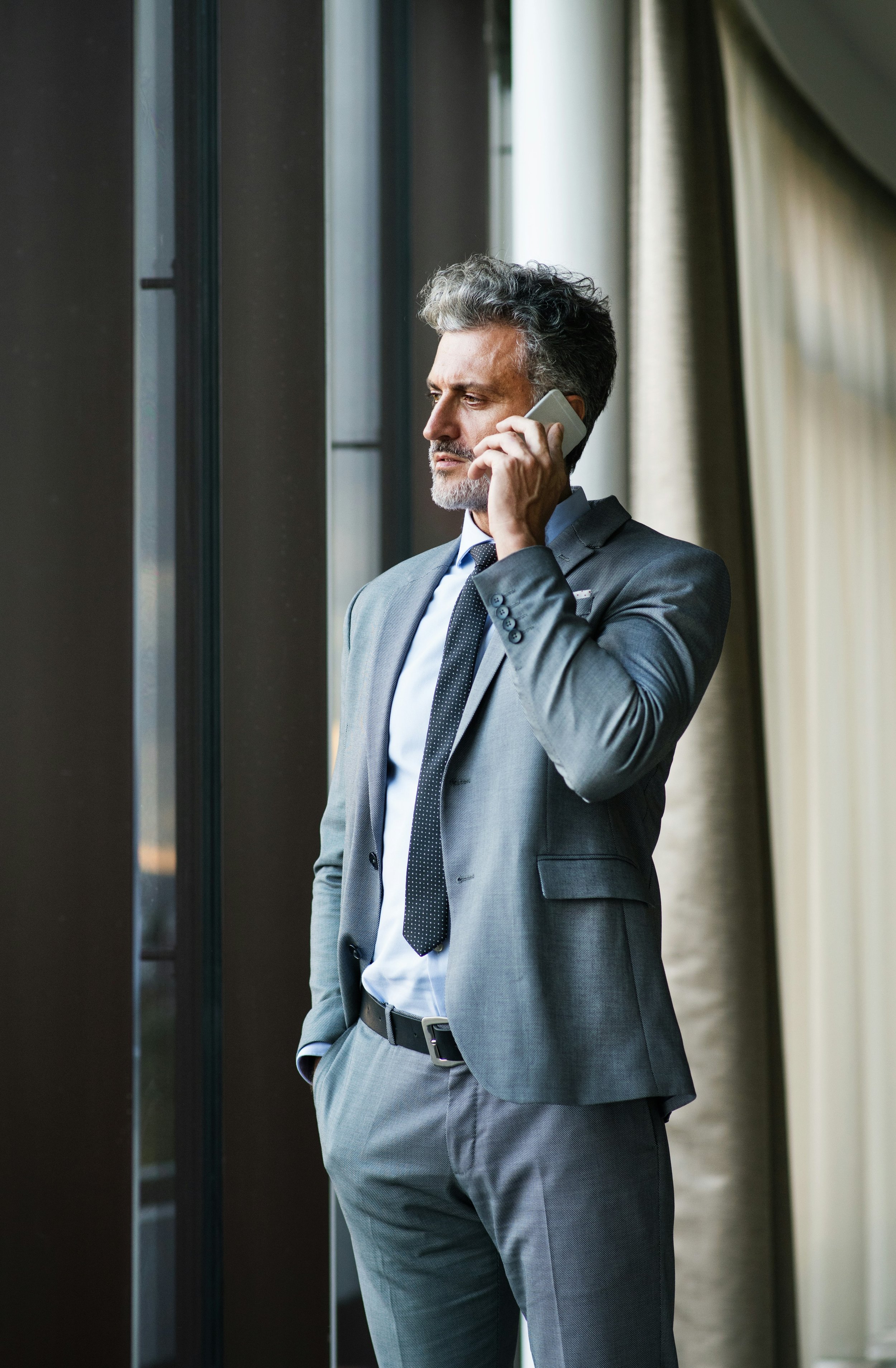A professional man in a grey suit and tie is standing by a window, talking on his cell phone in a modern office setting.
