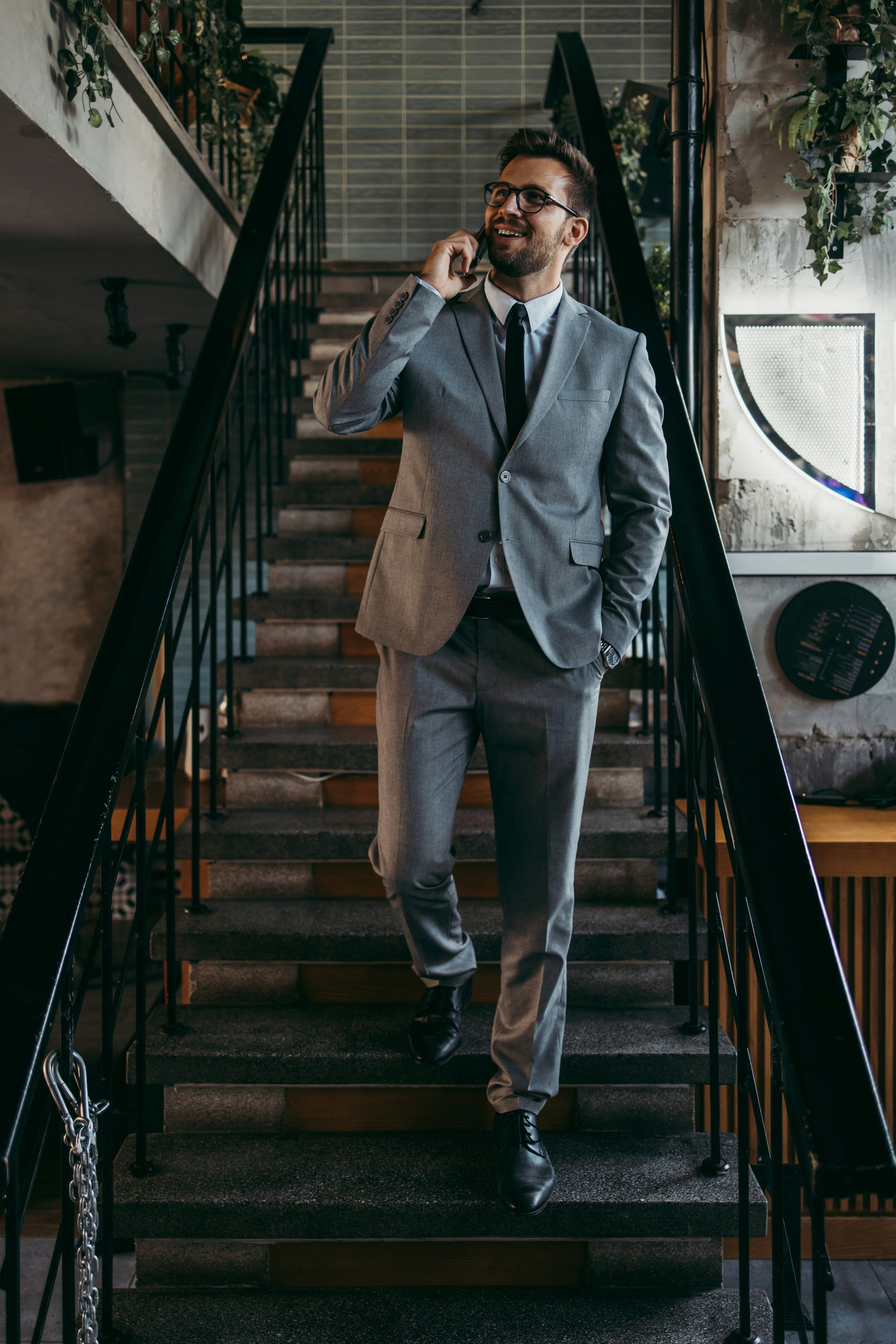 A man in a gray suit, black shoes, and glasses is walking down a staircase while talking on a cellphone, smiling and looking to the side.