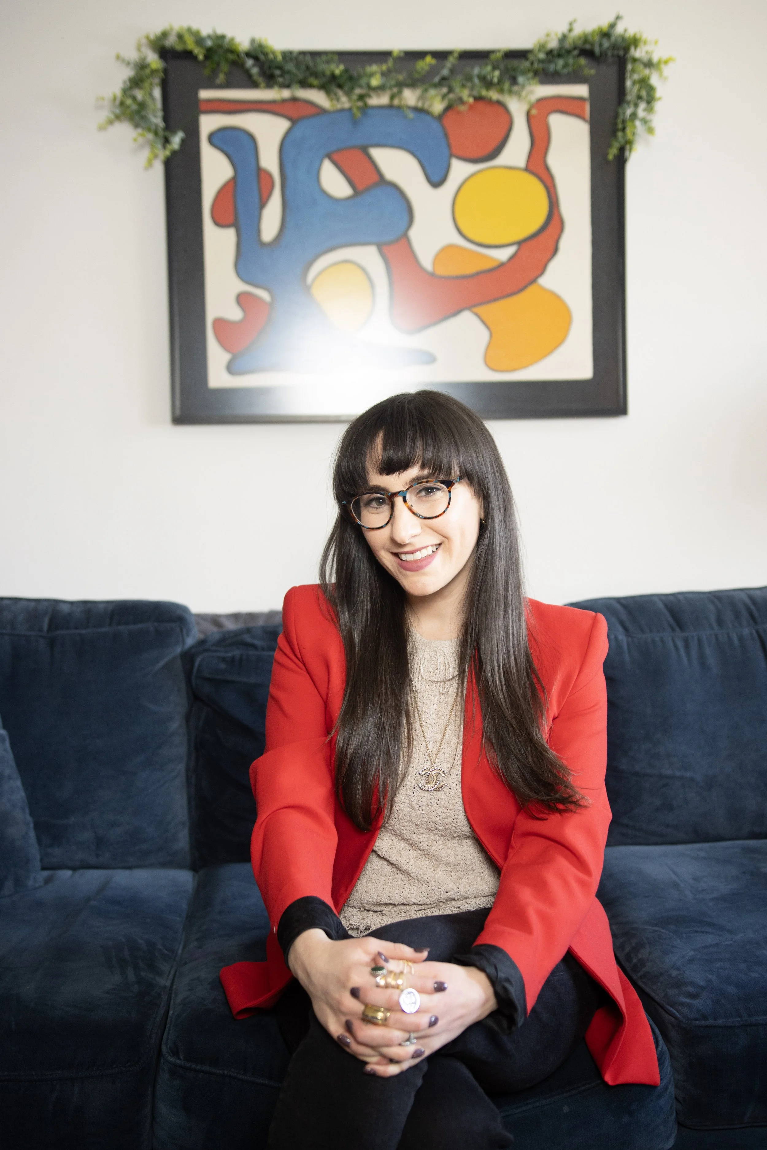 A woman with long dark hair and glasses wearing a red blazer and beige top sitting on a dark blue sofa. Behind her is an abstract painting with colorful shapes, framed and decorated with green foliage.