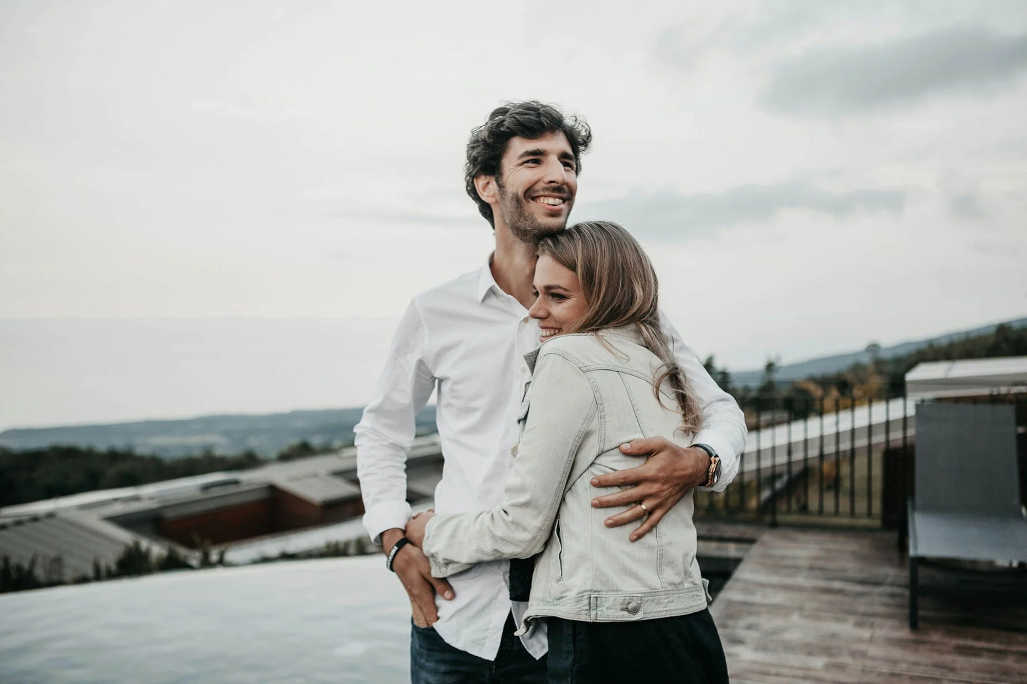 A man and woman hugging outdoors on a cloudy day, with a railing and rooftops in the background.