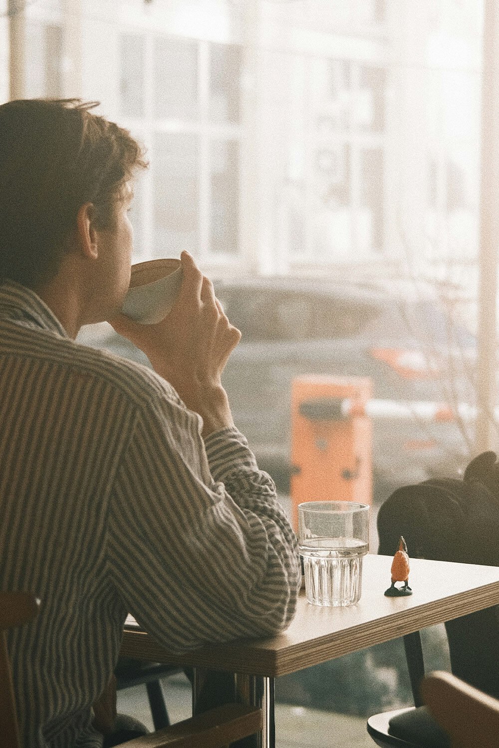 A man in a striped shirt drinks coffee at a wooden table in a coffee shop, with a glass of water and a small figurine on the table, near a large window with bright natural light.