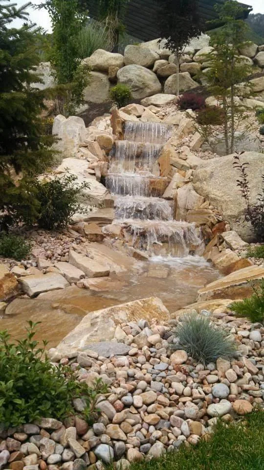 Small waterfall flowing over rocks surrounded by plants and pebbles in a landscaped garden.