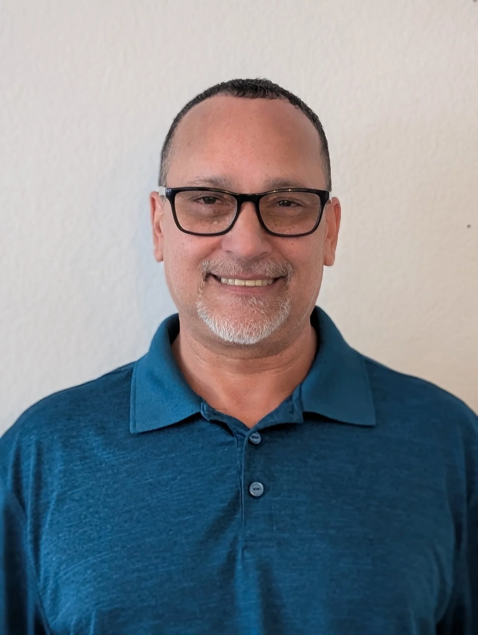 A man smiling and wearing glasses, a blue collared shirt, standing in front of a plain light-colored wall.