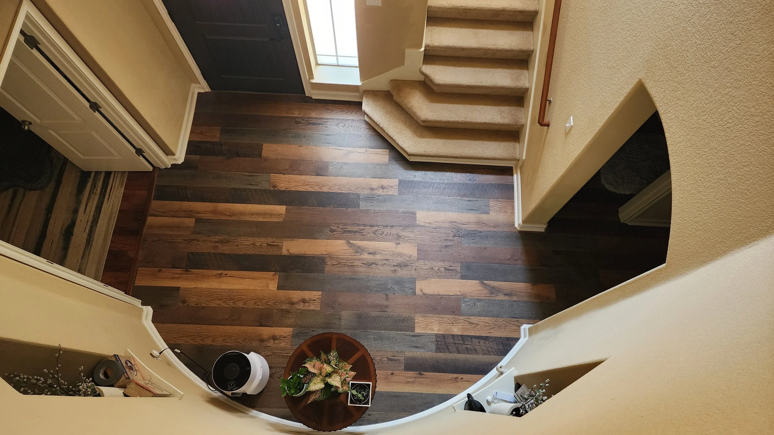 Interior view of a home entryway with hardwood floors, stairs with carpeted steps, a small round table with a flower arrangement, and a black front door.