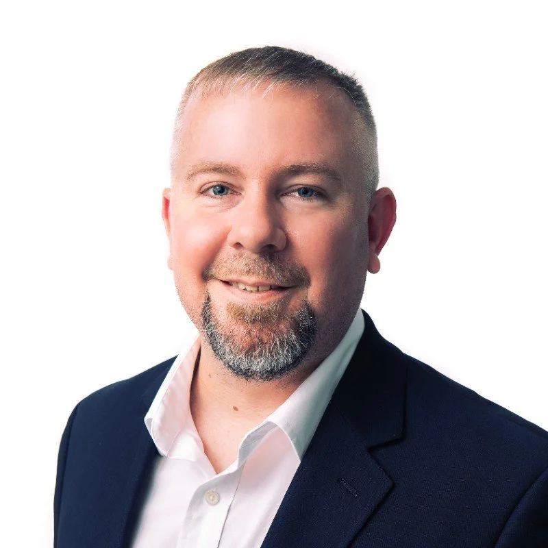 A professional headshot of a man with short blond hair, a goatee, wearing a white collared shirt and a navy blue blazer, smiling against a white background.