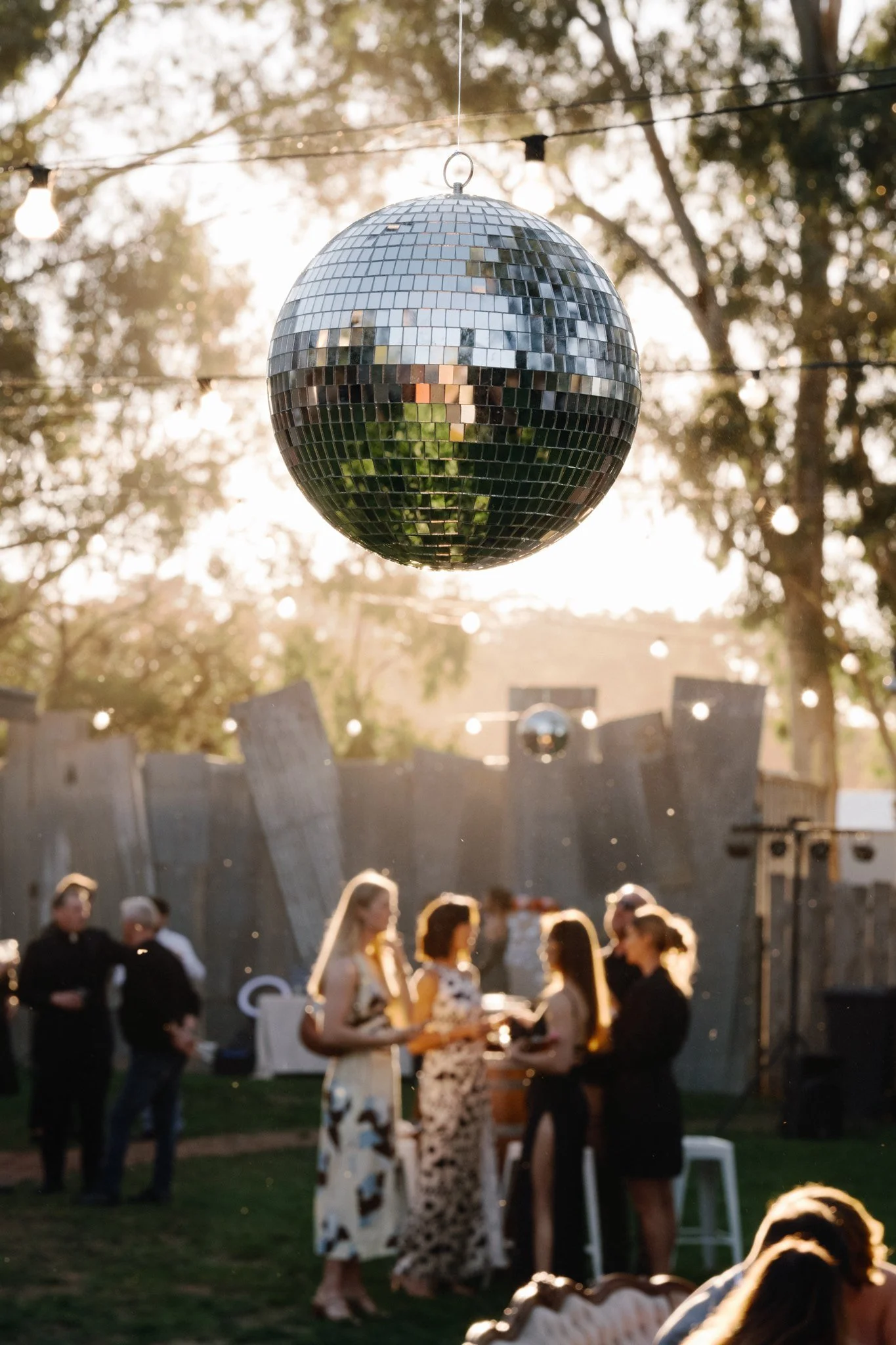 Disco ball hanging outdoors at a gathering or party, with people mingling and chatting in the background