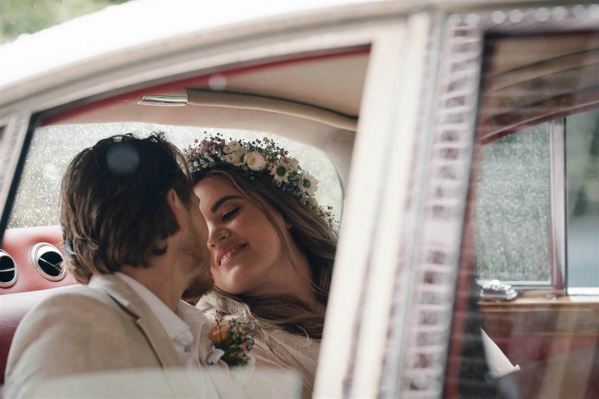 A bride and groom sharing a joyful kiss inside a vintage car, with the bride wearing a floral crown.