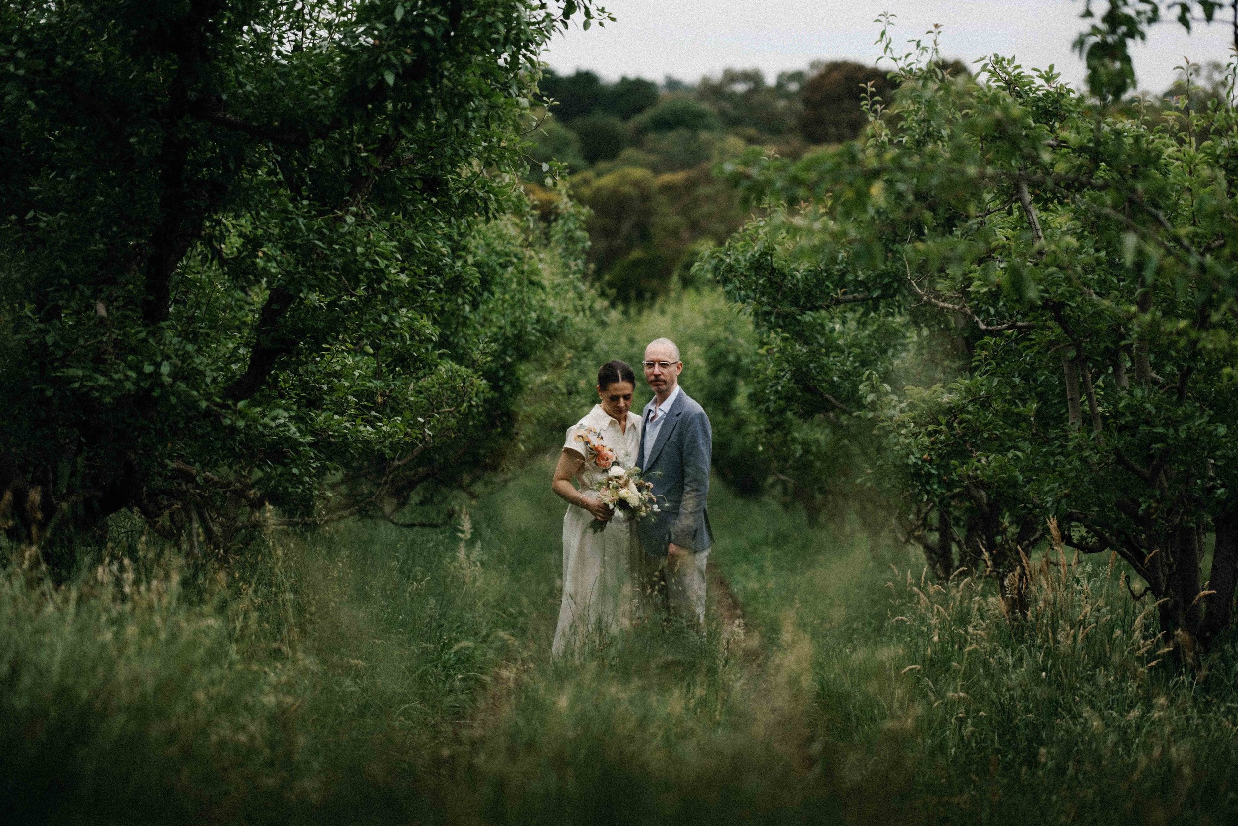 A couple standing in an orchard surrounded by trees, with the woman holding a bouquet of flowers and both dressed in semi-formal attire.