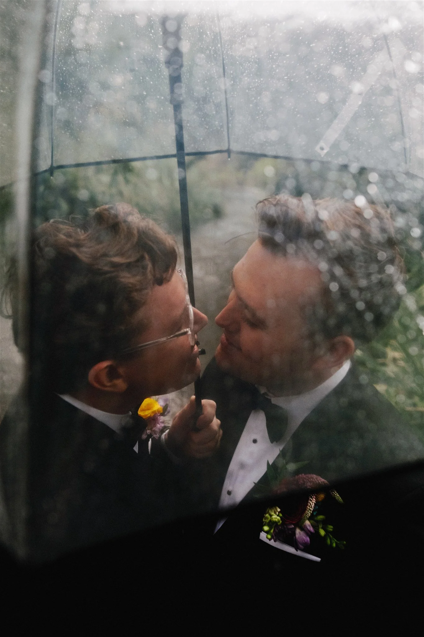 Two men in tuxedos sharing a close moment, viewed through a rain-covered glass umbrella.