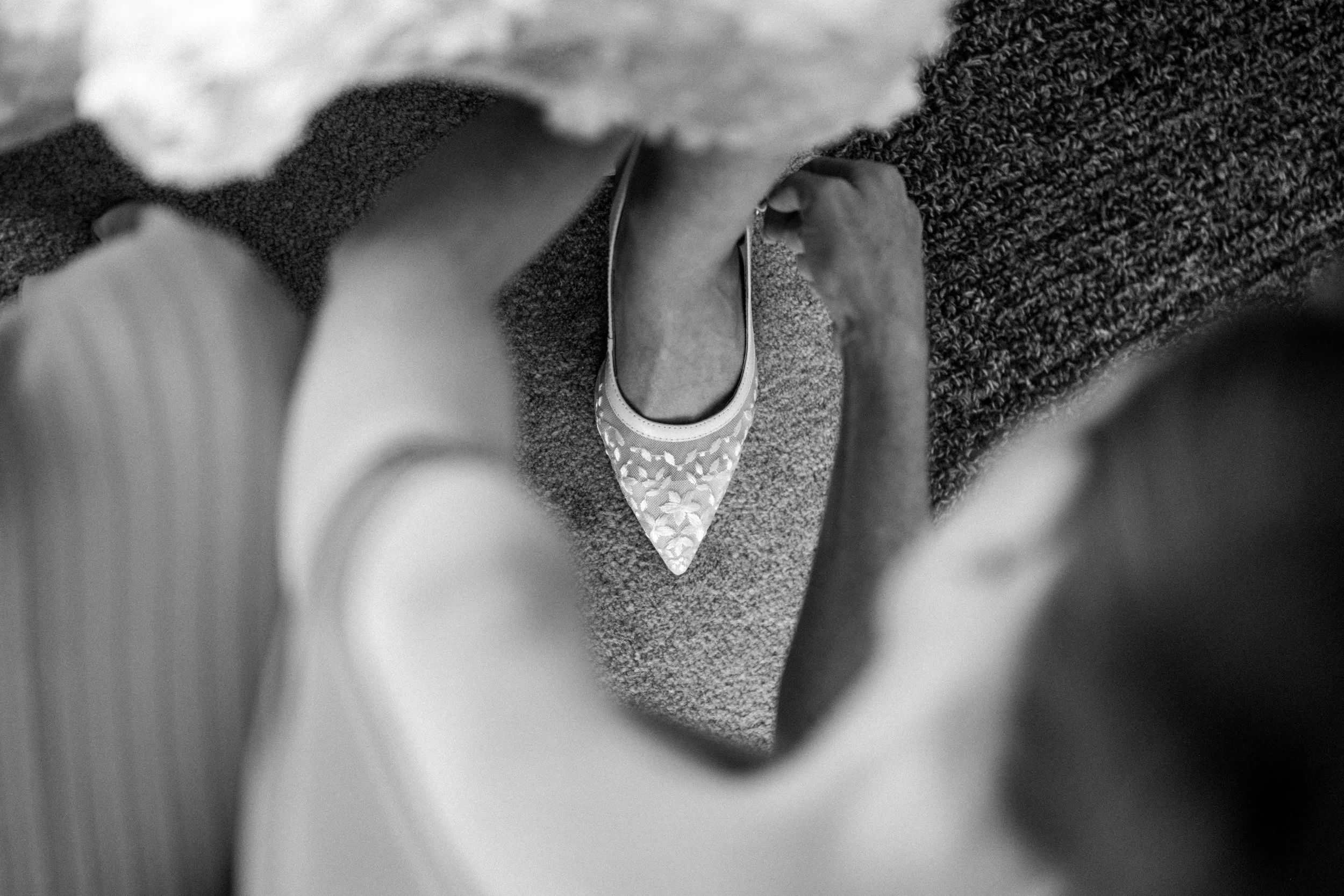 Black and white photo of a person tying the laces of a pointed-toe flat shoe with a floral pattern, viewed from above.