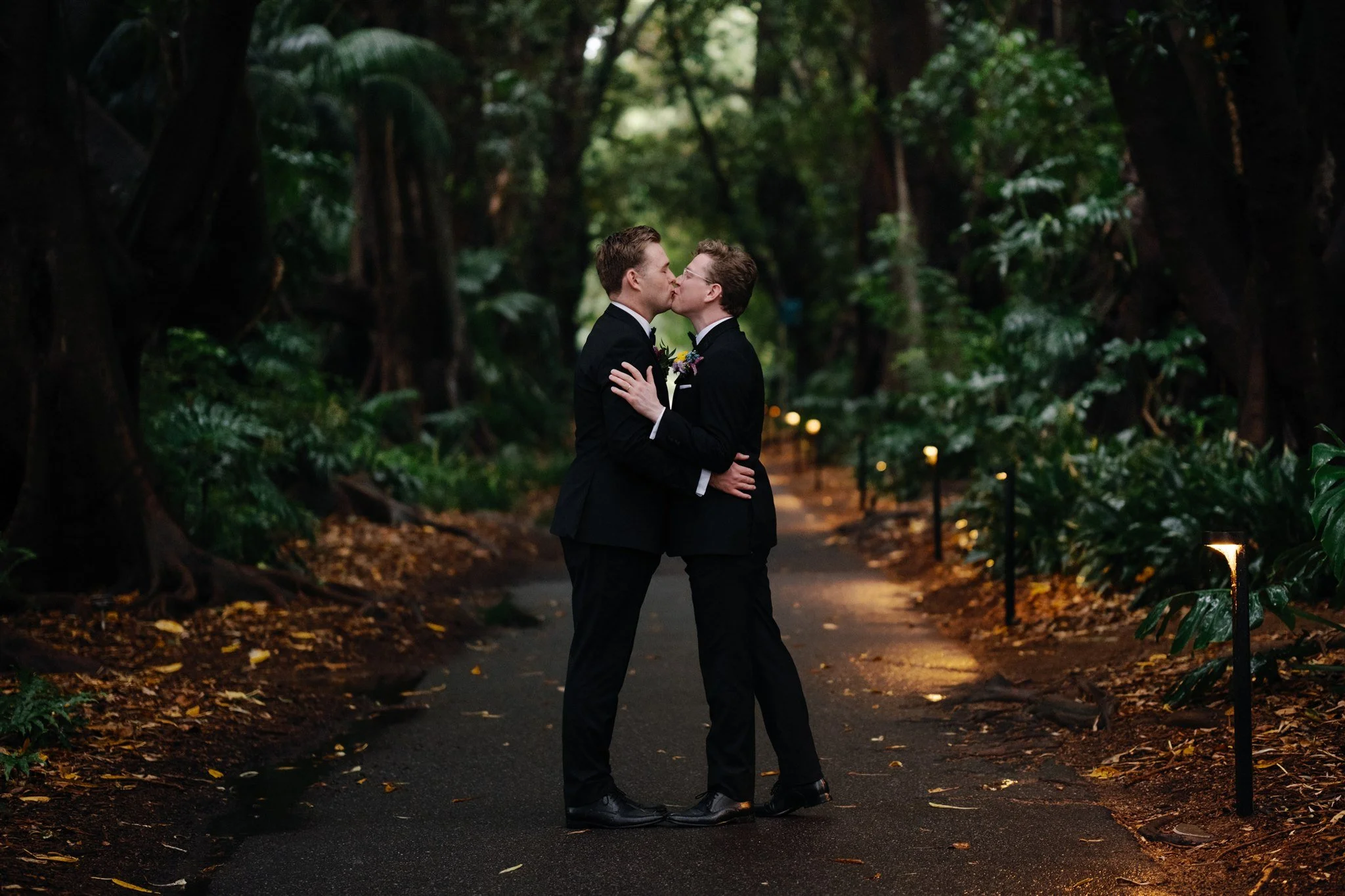 Two men in black suits sharing a kiss in a lush green forest with a pathway lit by small lights.