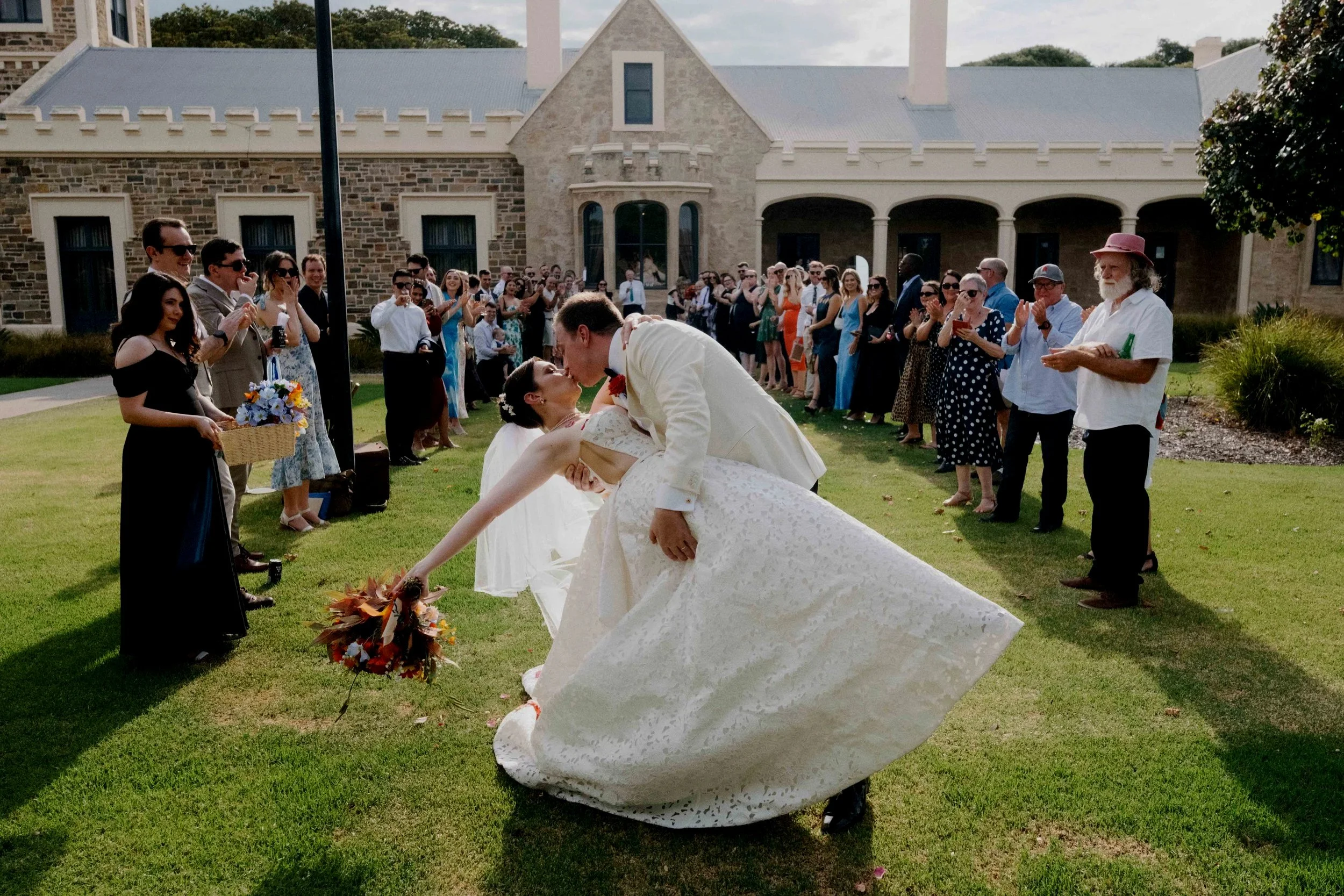 A bride and groom kiss as the groom dips the bride during their wedding celebration on the lawn of a large stone building, with wedding guests watching and clapping in the background.