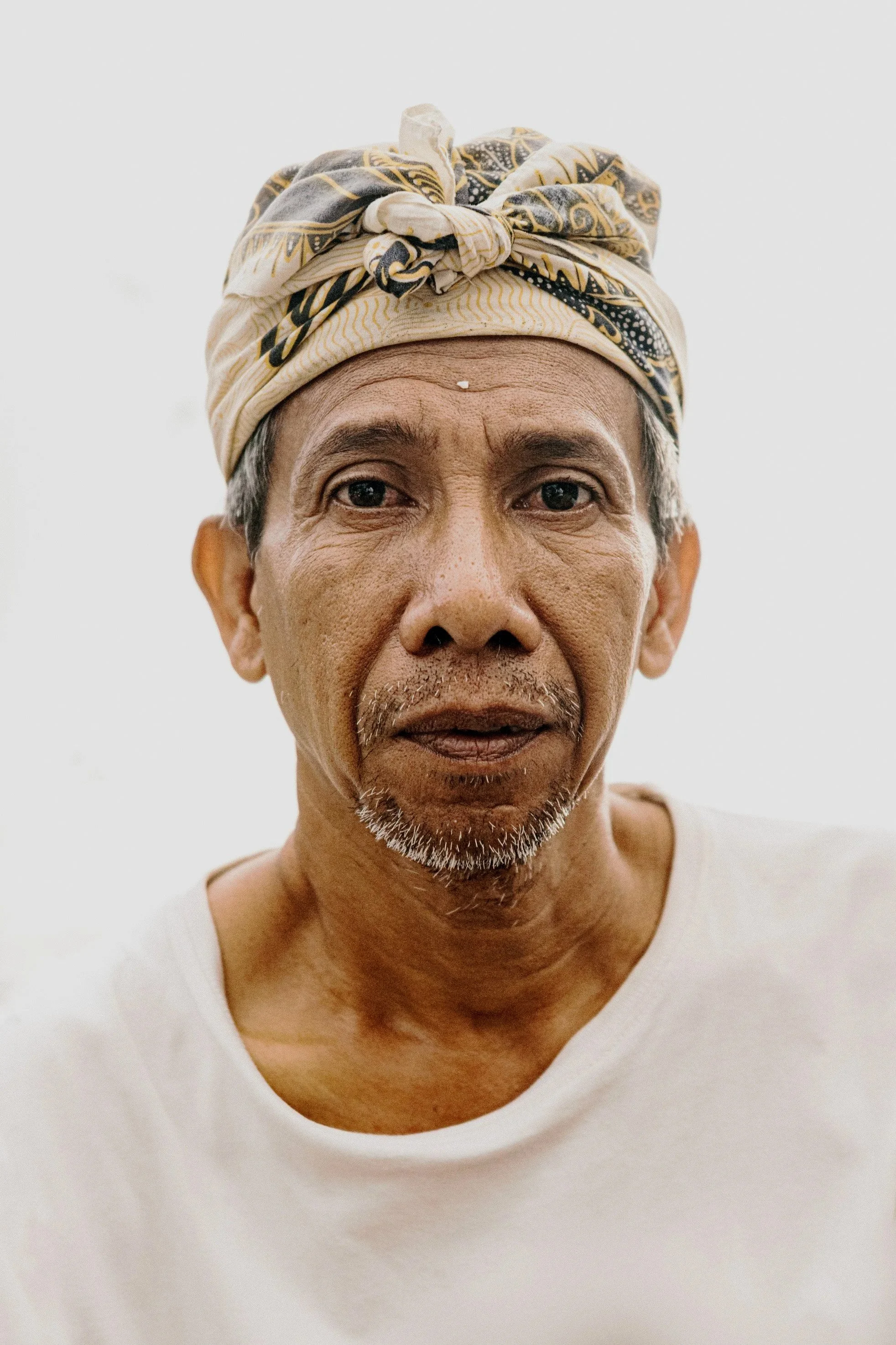 Close-up of an elderly man wearing a beige and black patterned headscarf and a white shirt, with a neutral expression.