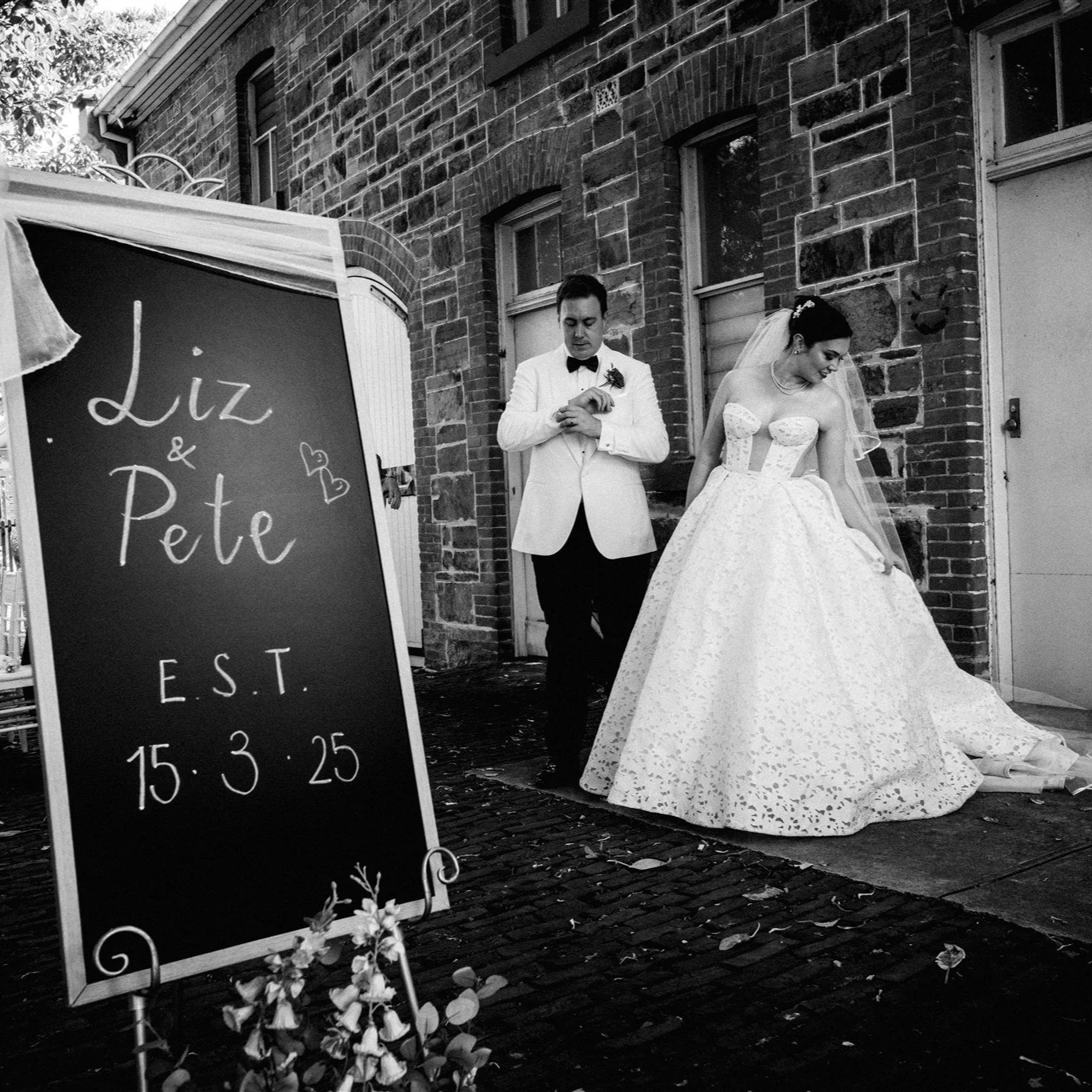 A wedding couple standing outside a brick building, with a sign that reads "Liz & Pete, EST. 15.3.25."