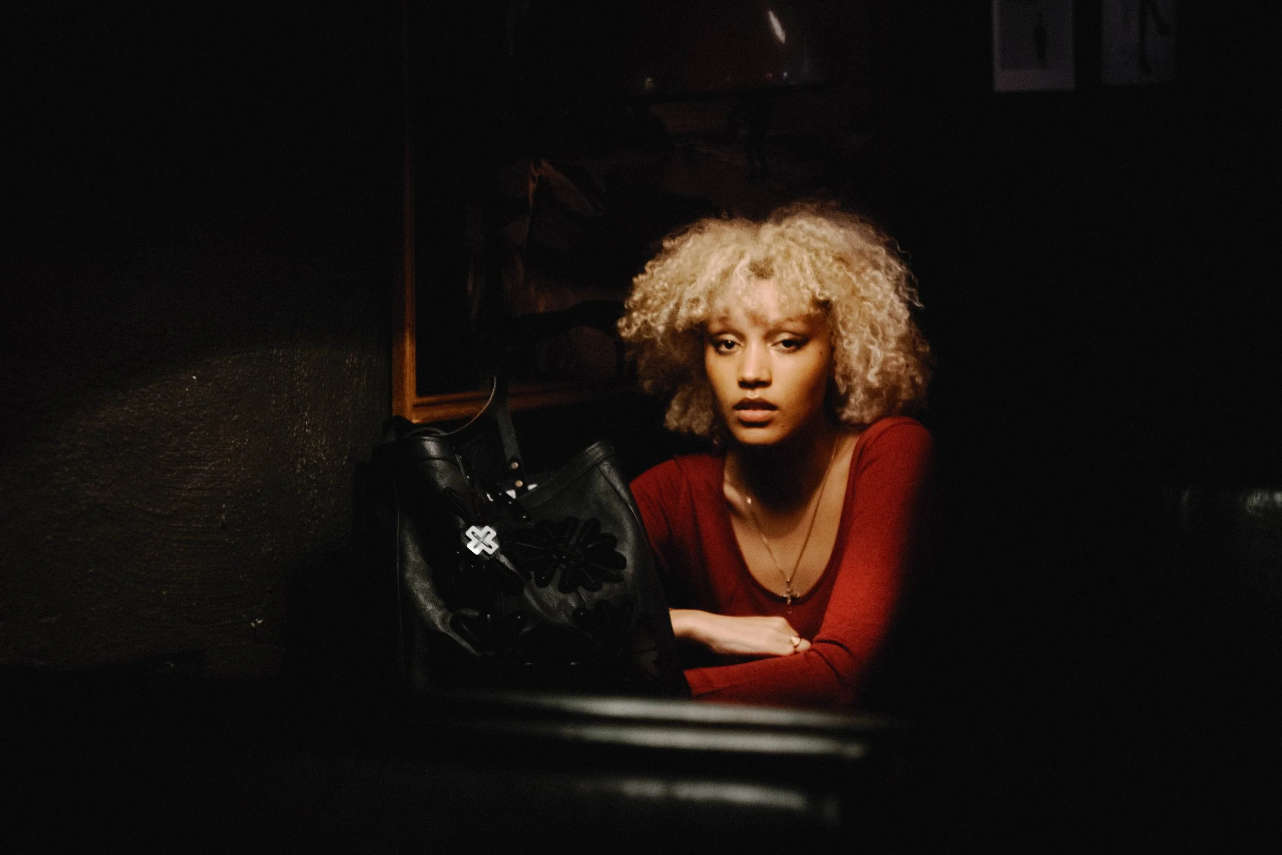 A woman with curly blonde hair wearing a red top sitting in a dark room. She is looking towards the camera with a serious expression, and there is a black purse in front of her.