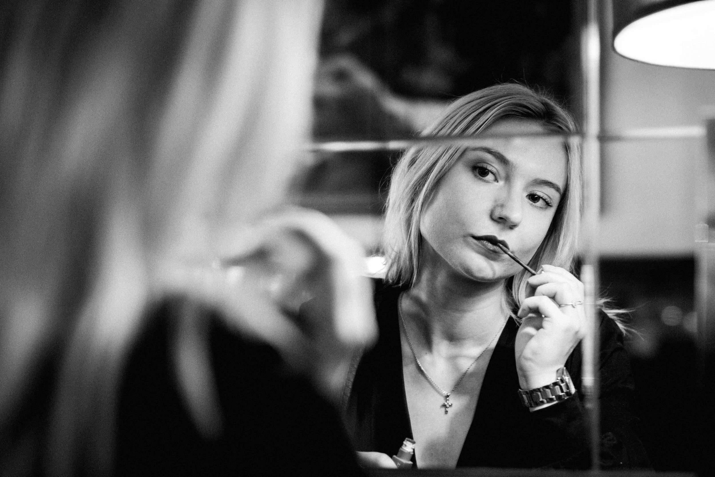 Black and white photo of a young woman with blonde hair looking at her reflection in a mirror, holding a lipstick or makeup applicator near her lips.