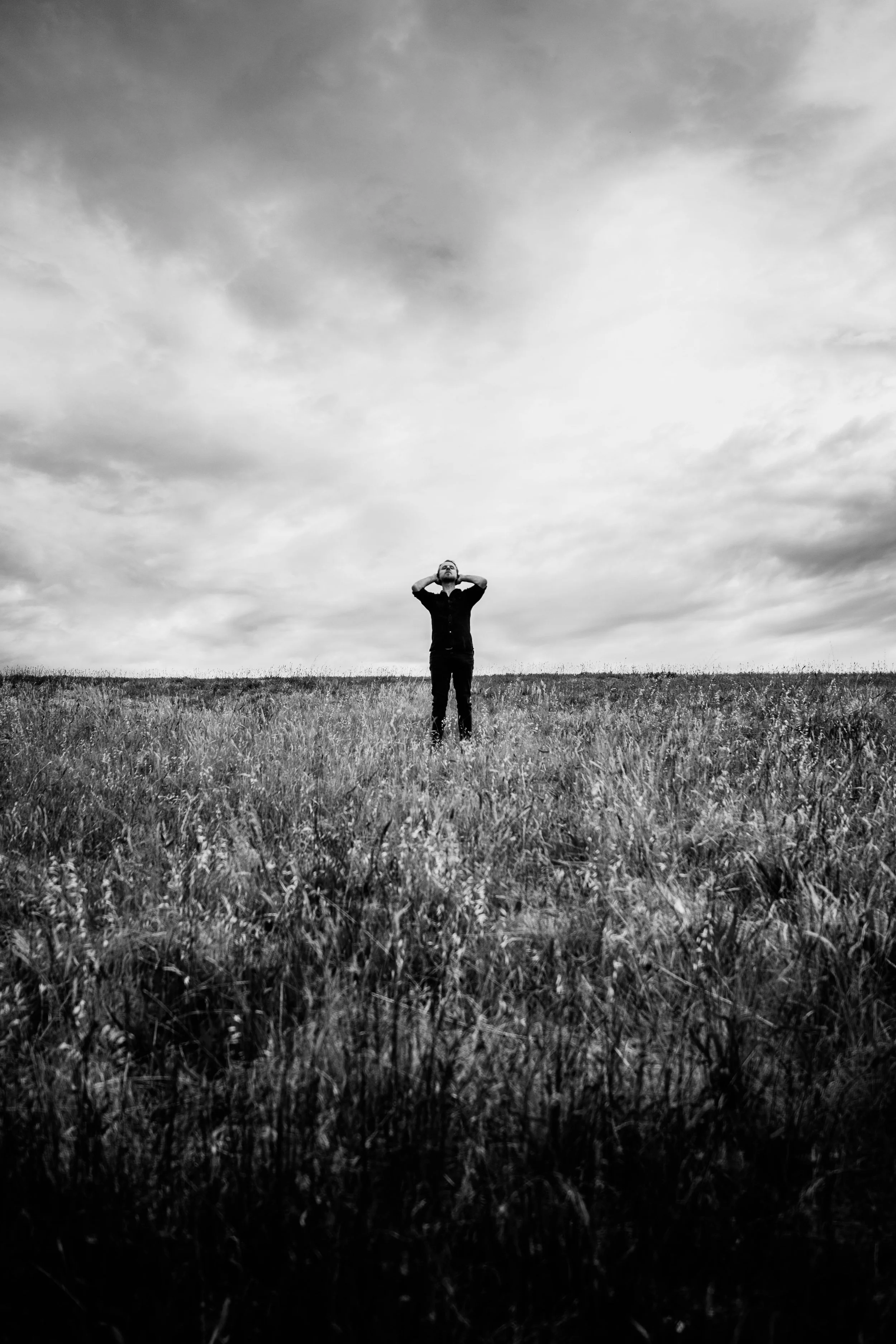 A person standing alone in a grassy field under cloudy sky, looking upwards with hands behind their head, in black and white.