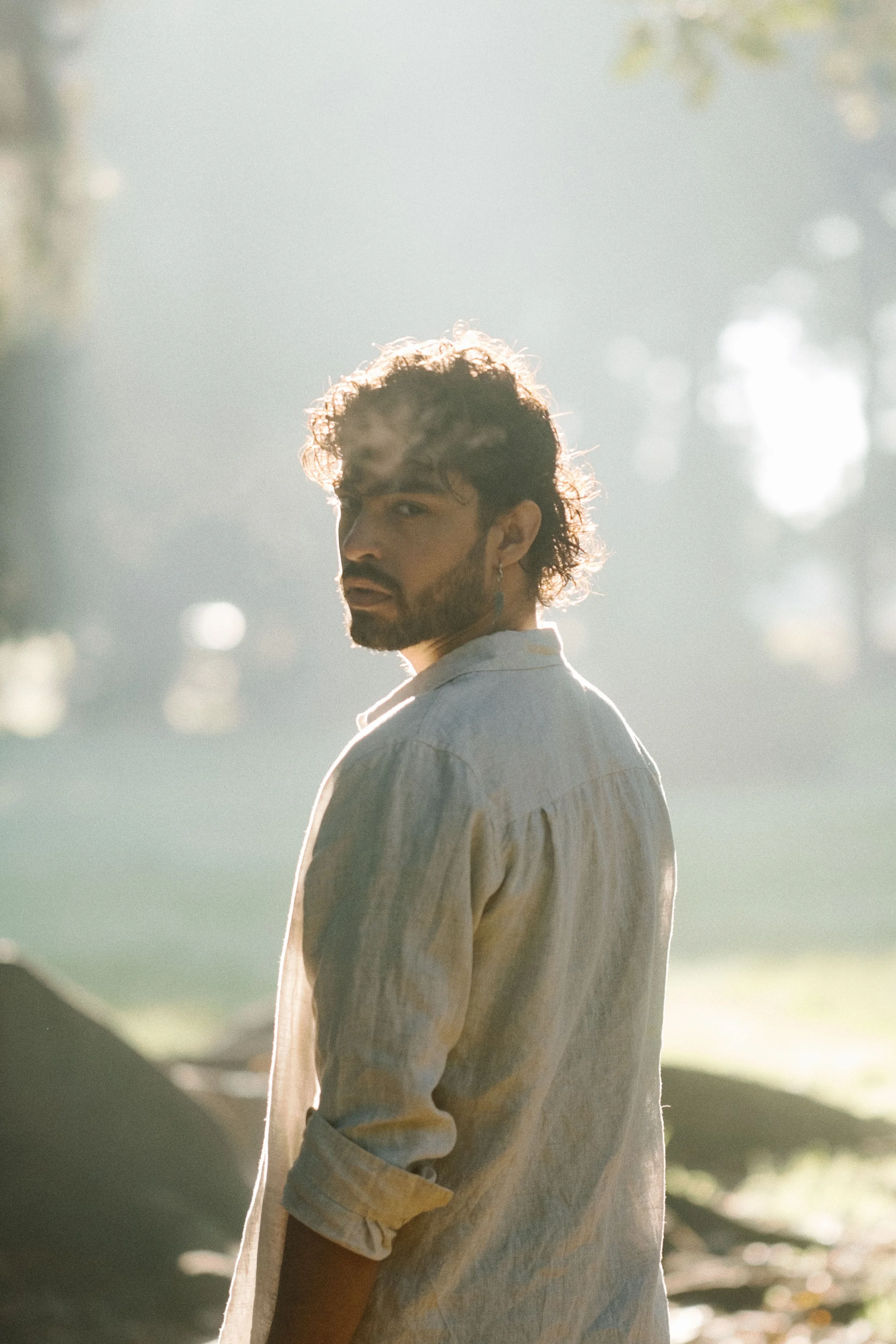 A man with curly hair and a beard wearing a light-colored shirt, standing outdoors with sunlight filtering through trees in the background.