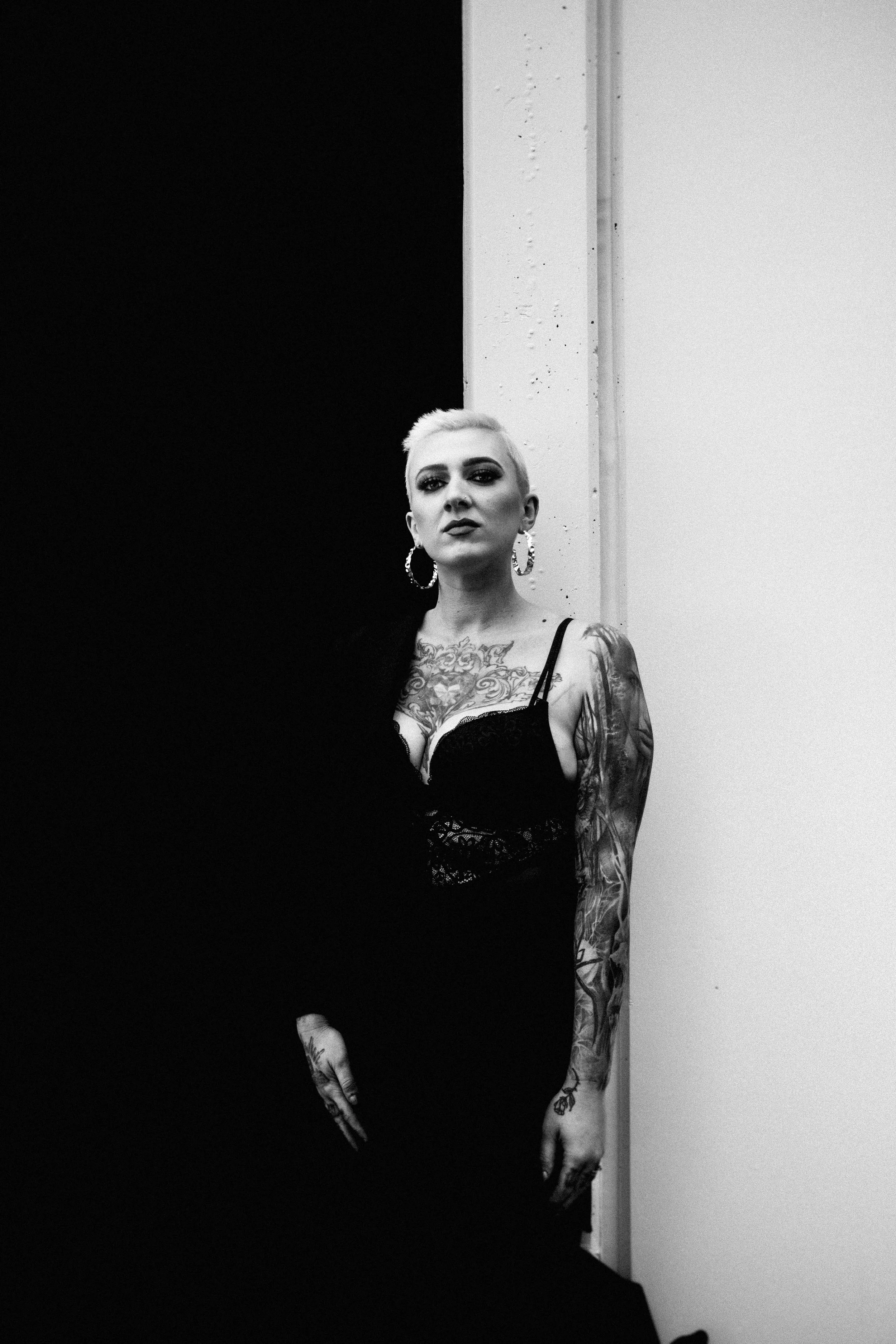 A woman with platinum blonde hair and tattoos on her arms, wearing a black lace top and large hoop earrings, standing against a wall with a contrasting dark and light background.
