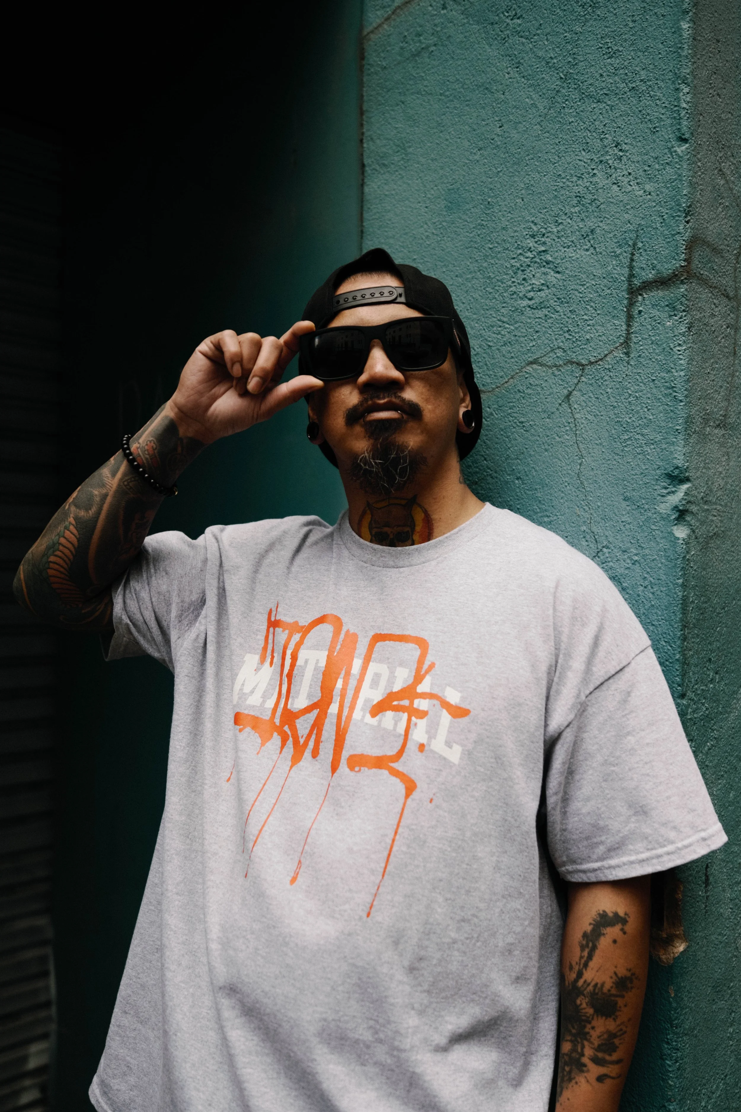 Man with tattoos, sunglasses, and a black cap adjusting sunglasses, standing against a textured teal wall.