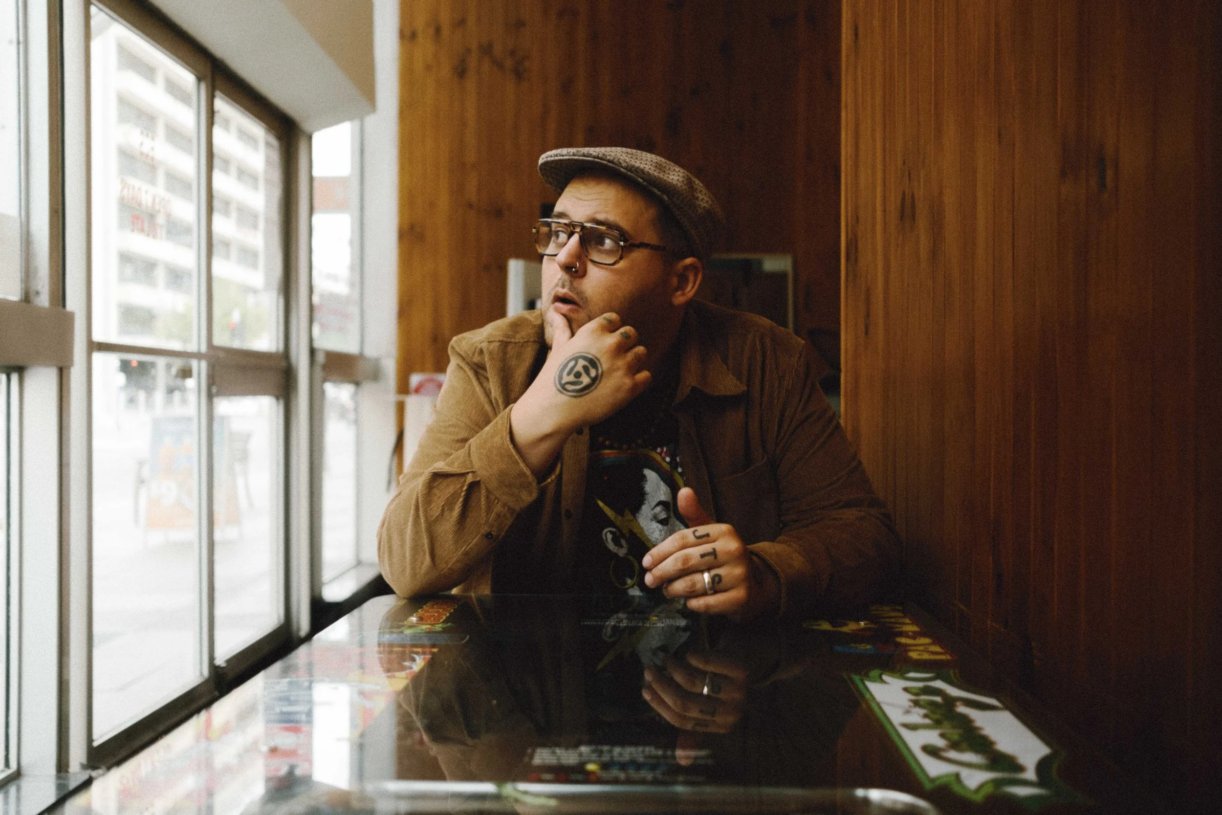 A man wearing glasses, a cap, and a brown jacket sitting at a table inside a room with wood-paneled walls. The man has tattoos on his fingers and his hand on his chin, looking to the side with a pensive expression.