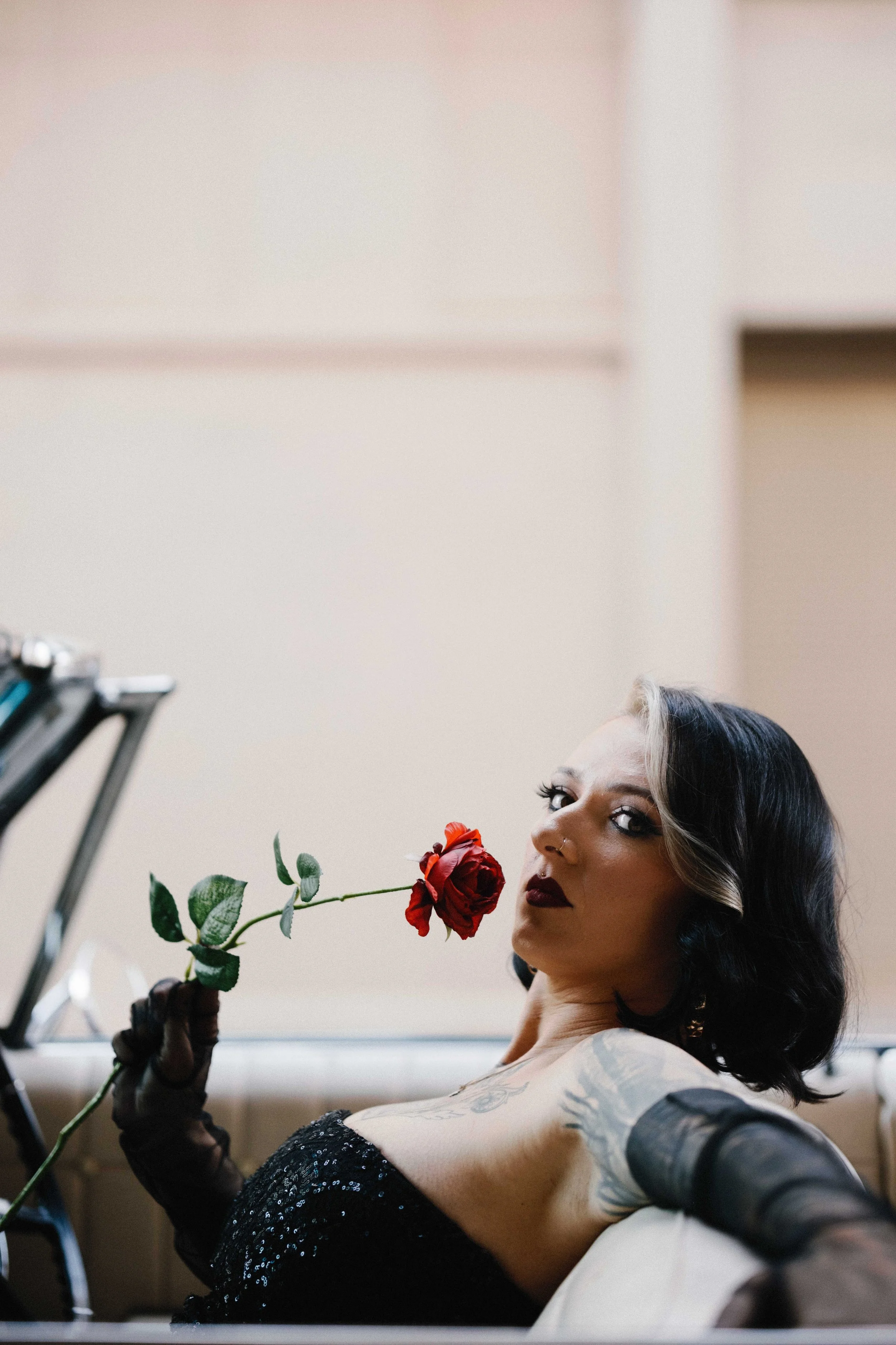 A woman with black and blonde hair and tattoos on her arm is holding a red rose in her hand. She is wearing dark lipstick and a black, sparkly off-shoulder top, and is sitting in a room with beige walls.