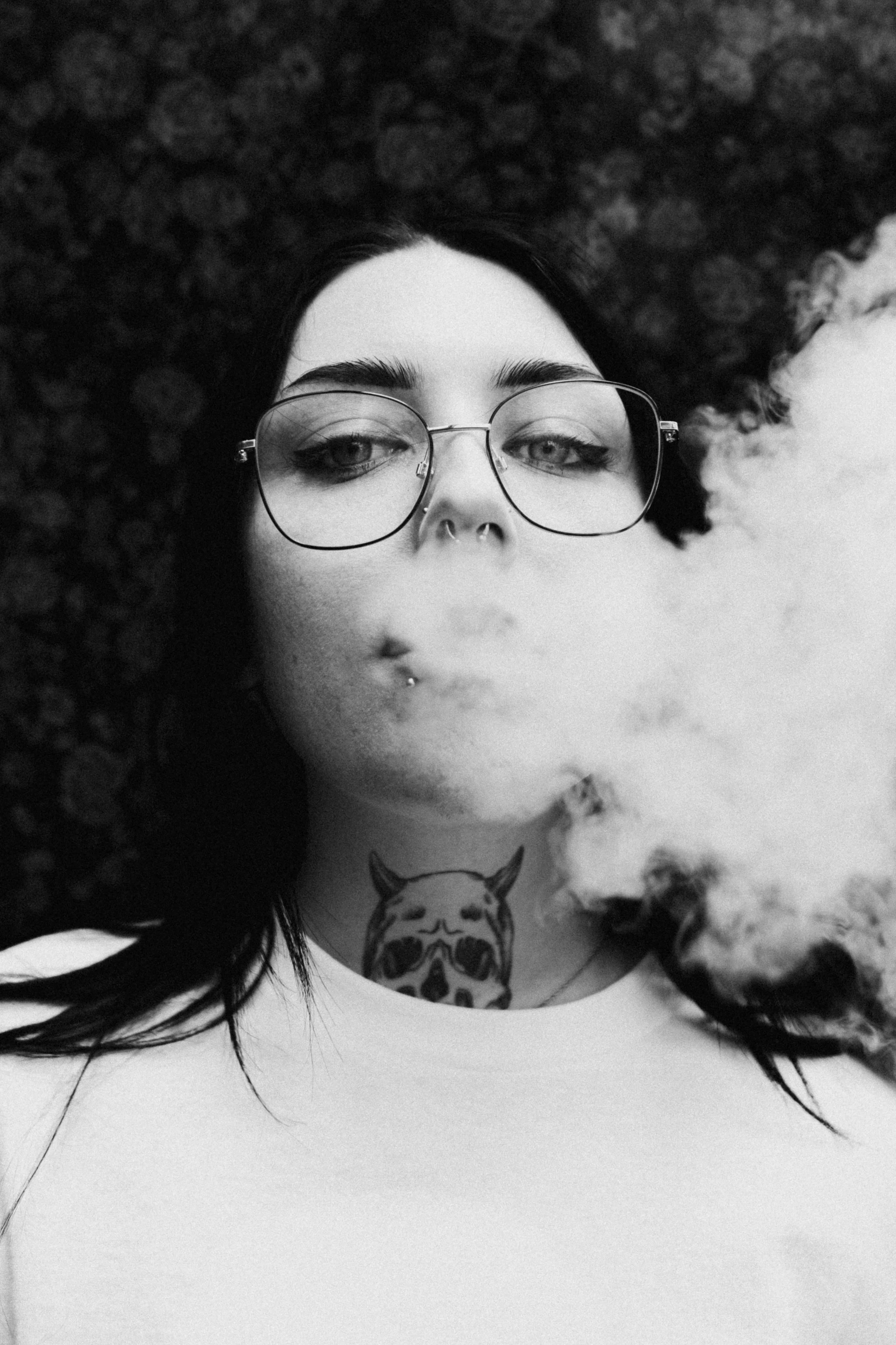 A black-and-white photo of a woman with dark hair, glasses, and septum piercing, lying on a pebble surface, exhaling vapor or smoke.