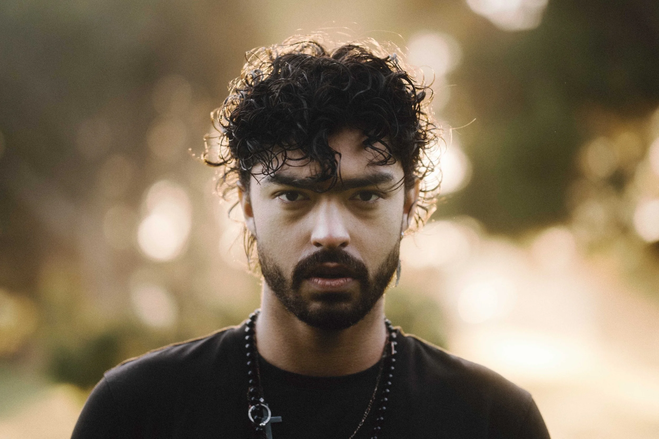 A man with curly dark hair and a beard looking directly into the camera outdoors during sunset, with bokeh light effects in the background.