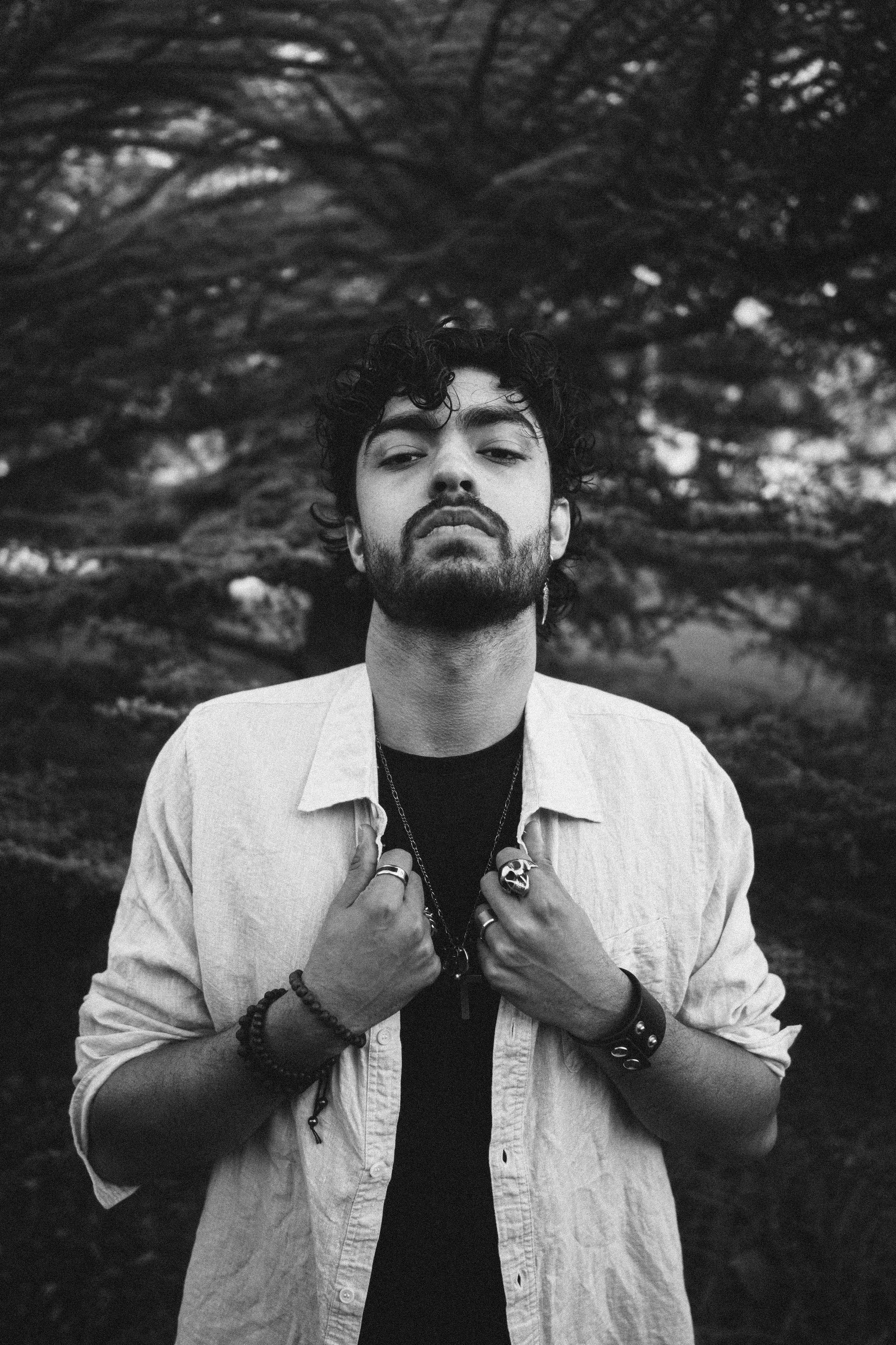 A black and white photo of a man with curly hair and a beard, wearing rings, bracelets, and necklaces, standing outdoors in front of trees, with a confident pose and serious expression.