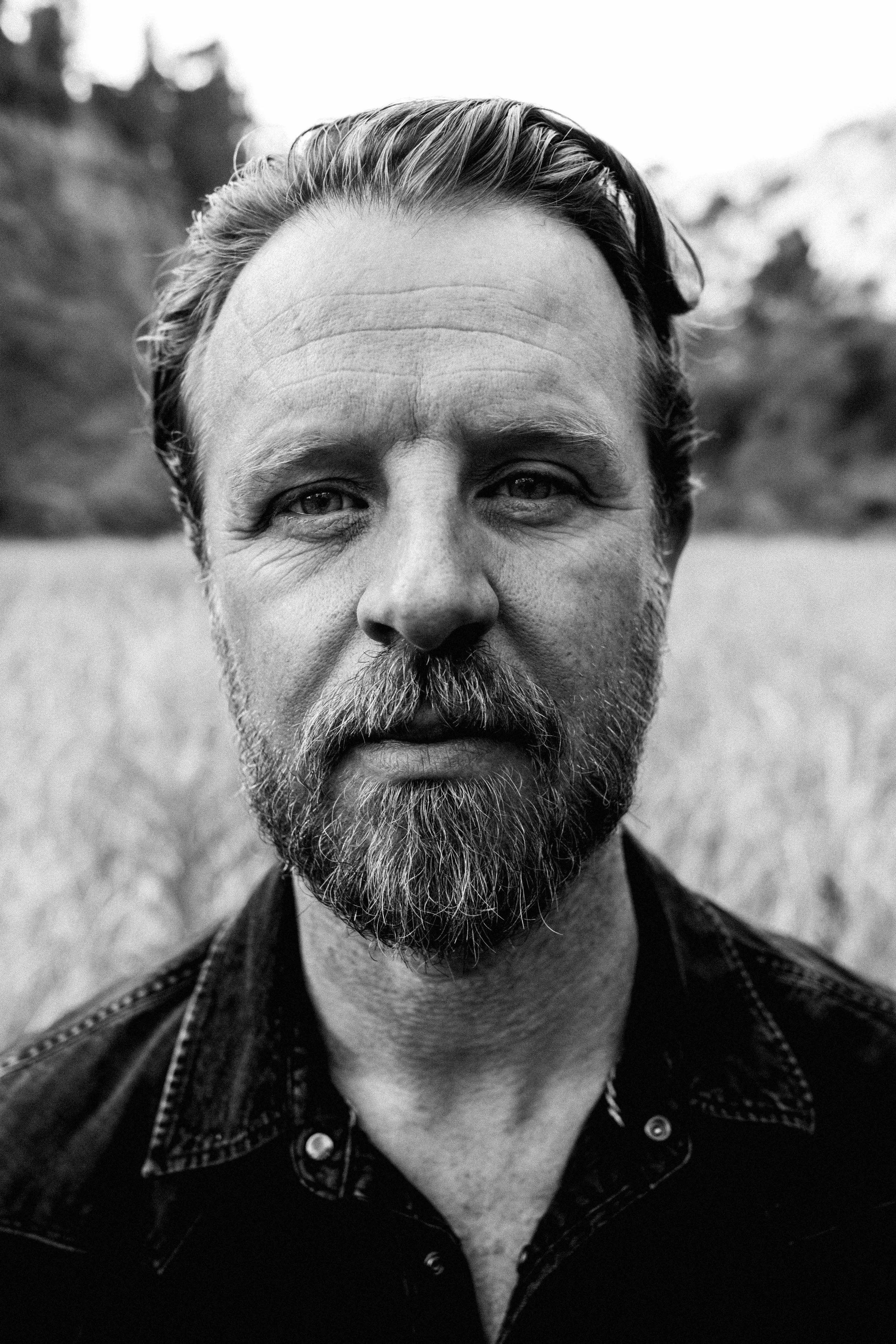Close-up black and white portrait of a bearded man outdoors, with a blurred natural background.
