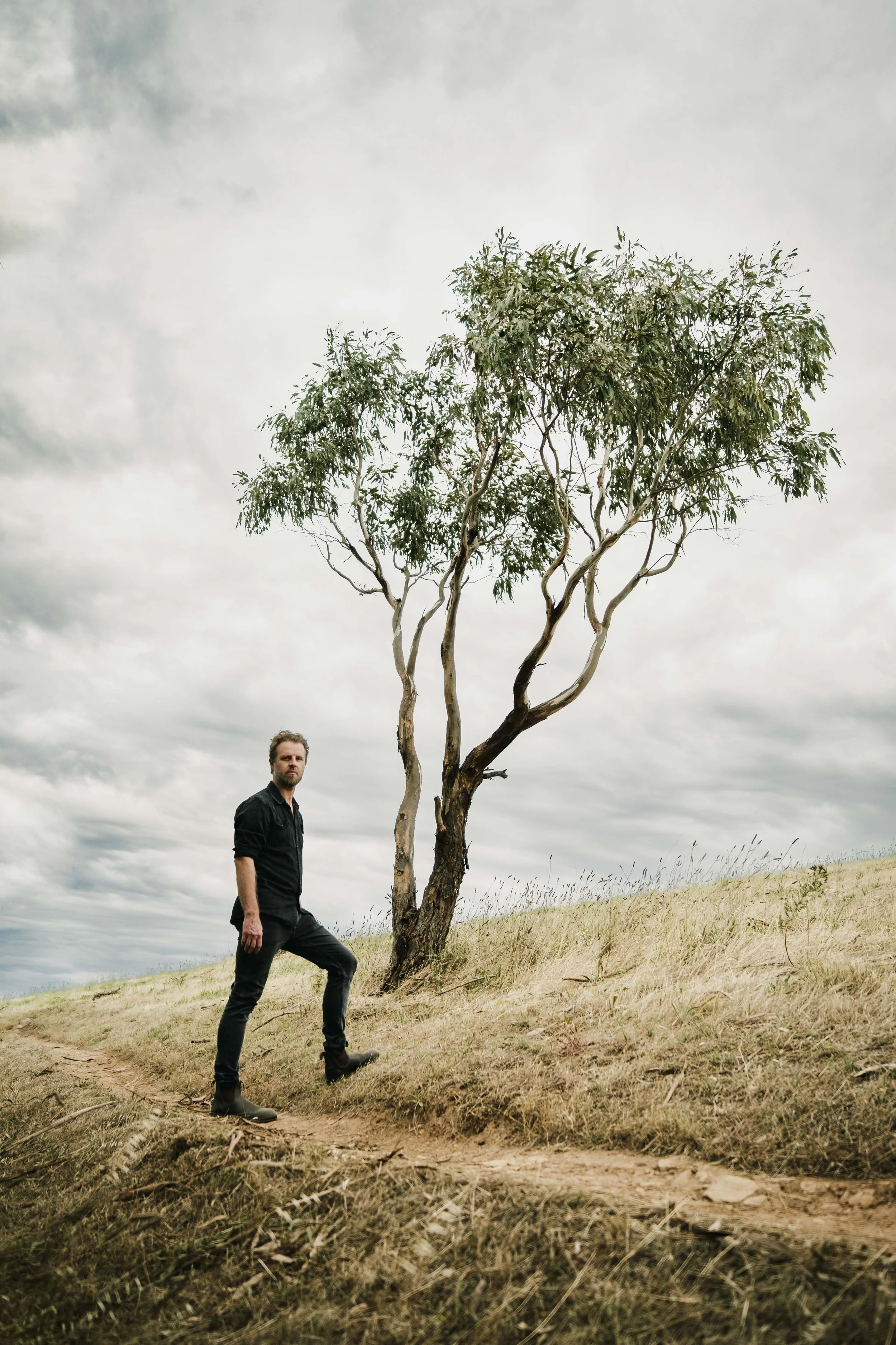 A man with a beard in black clothing standing on a dirt path on a hillside, near a leafless tree with sparse green leaves, under a cloudy sky.