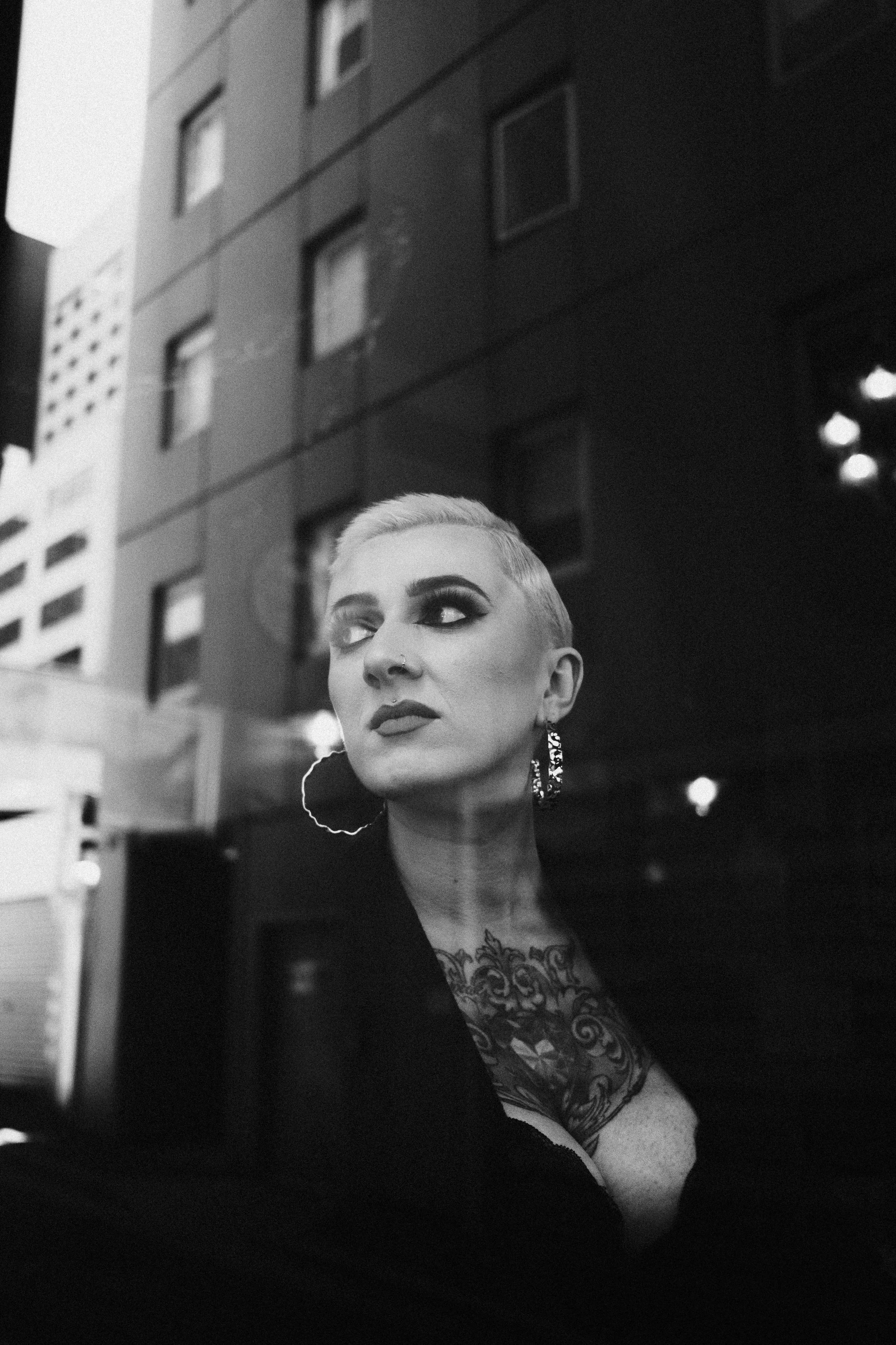 A black and white photo of a woman with short, platinum blonde hair and bold makeup, looking to the side. She has large earrings, visible tattoos on her chest, and is wearing a dark top. She is outdoors in an urban setting with a tall building in the