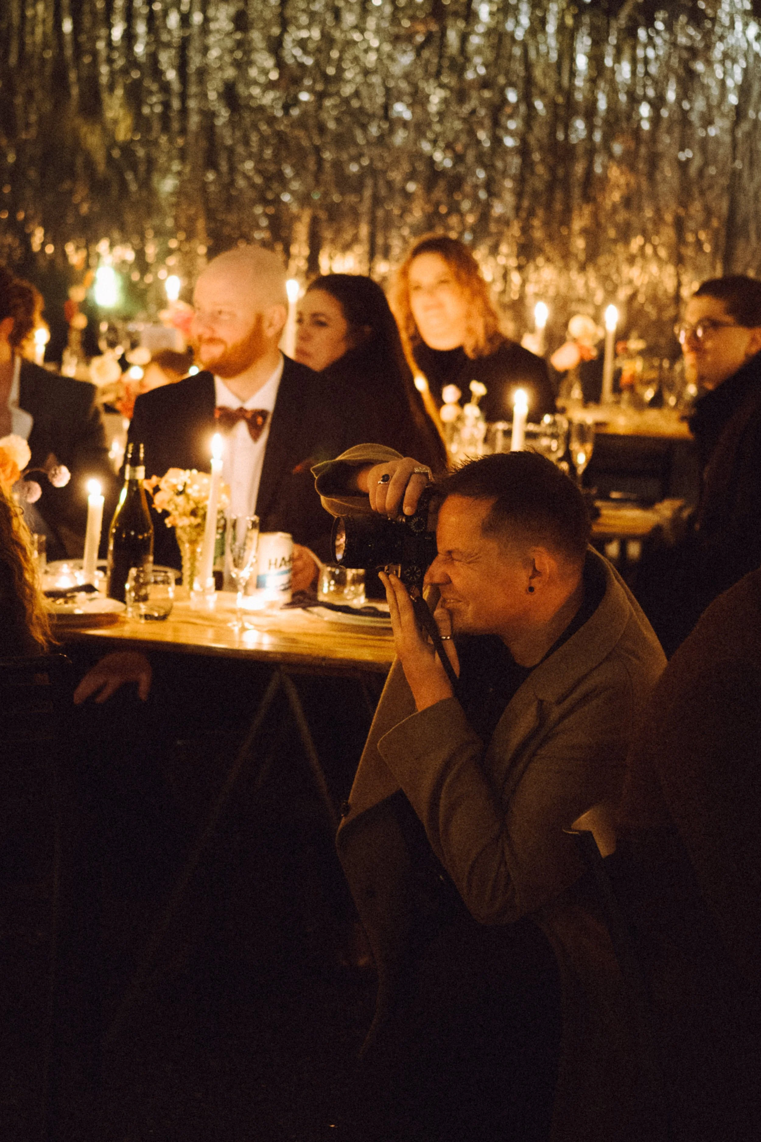 Photographer taking pictures of guests at a candle-lit indoor or outdoor event with a forest backdrop, dappled light, candles, flowers, wine, and drinks on tables.