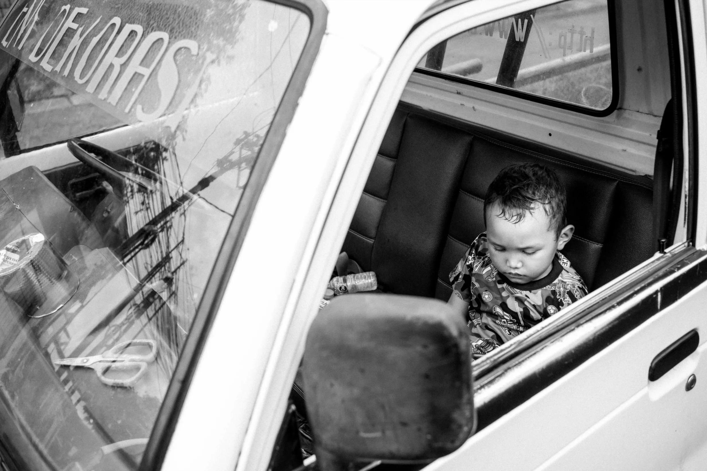A young boy sitting in the back seat of a vehicle, looking down. The vehicle appears to be an older model with manual controls and visible interior items like scissors and cans on the dashboard. The photo is in black and white, taken from outside thr