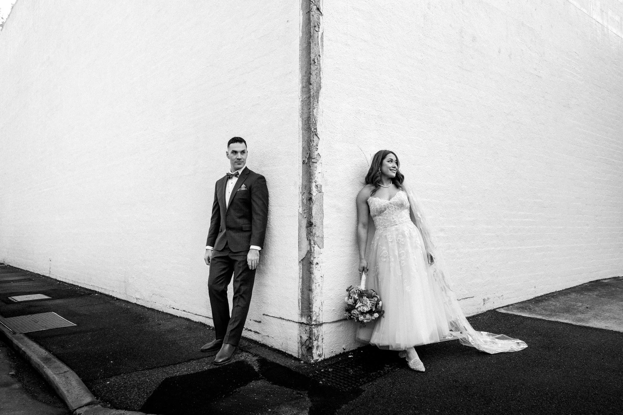 A black and white photo of a man in a tuxedo and a woman in a wedding dress leaning against opposite sides of a corner wall, facing away from each other, with the woman holding a bouquet of flowers.