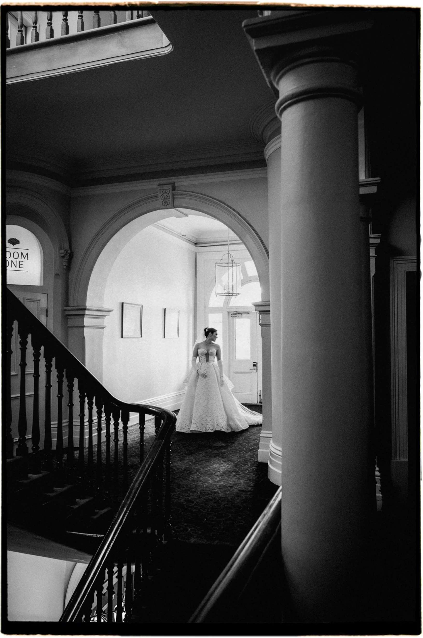 A woman in a wedding dress standing in a bright, elegant hallway with arches and large windows, black and white photo.