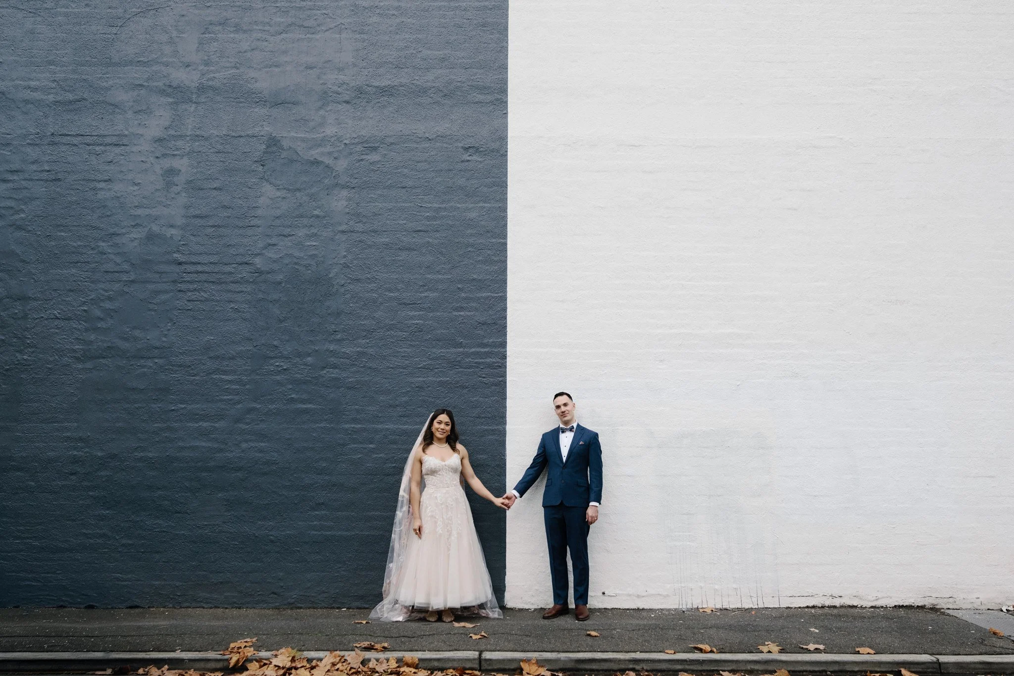 A bride and groom holding hands, standing in front of a split-colored wall with one side dark gray and the other white, outdoors with fallen leaves on the ground.