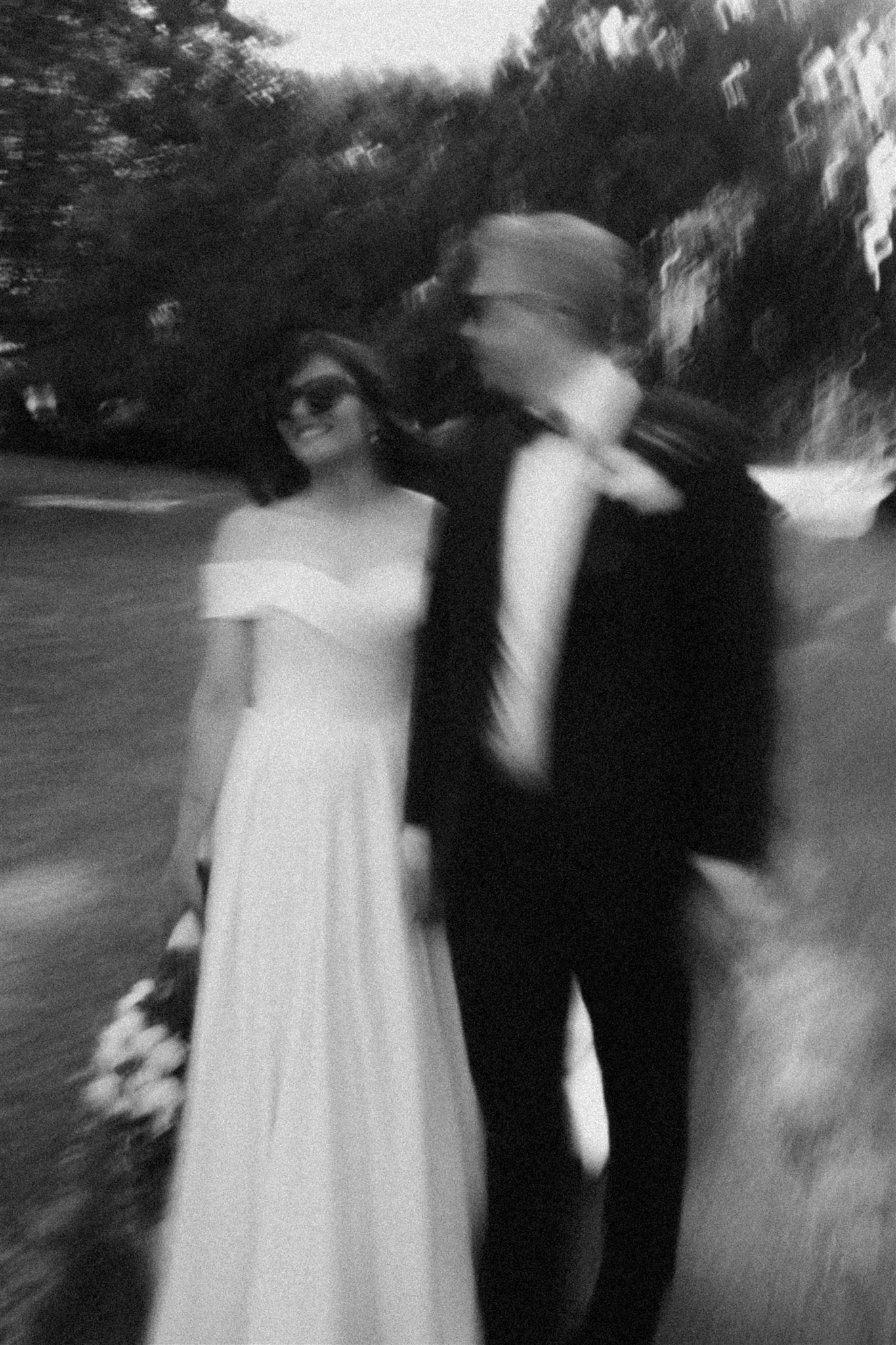 A black and white photo of a woman in an off-shoulder dress holding a bouquet, standing next to a man in a suit with a hat, outdoors with trees in the background.